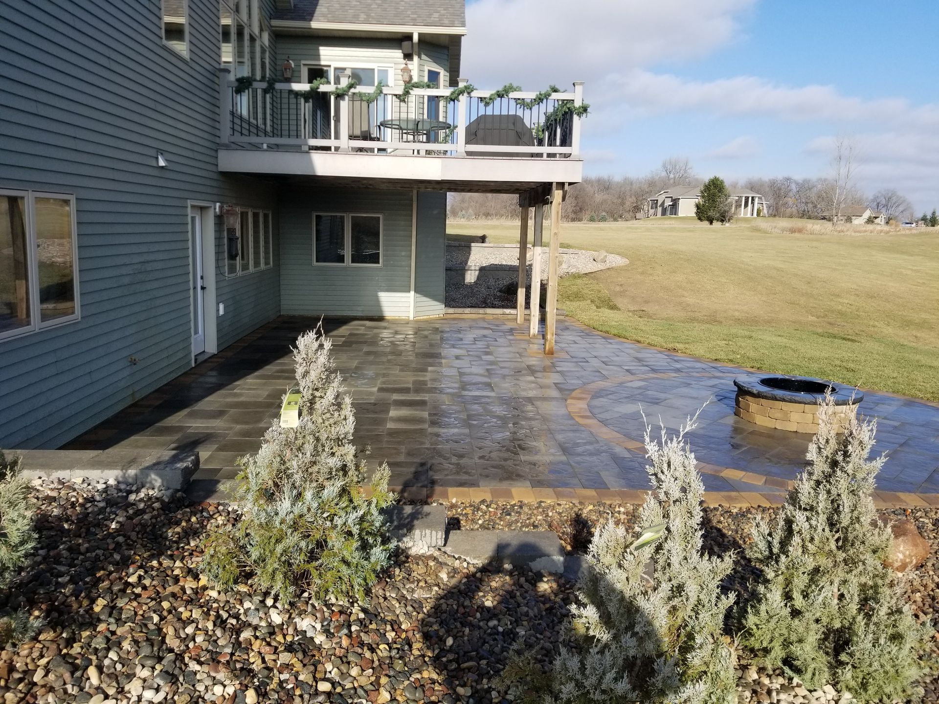 A blue house with a raised wooden deck over a stone patio featuring a circular fire pit, overlooking an open grassy field.