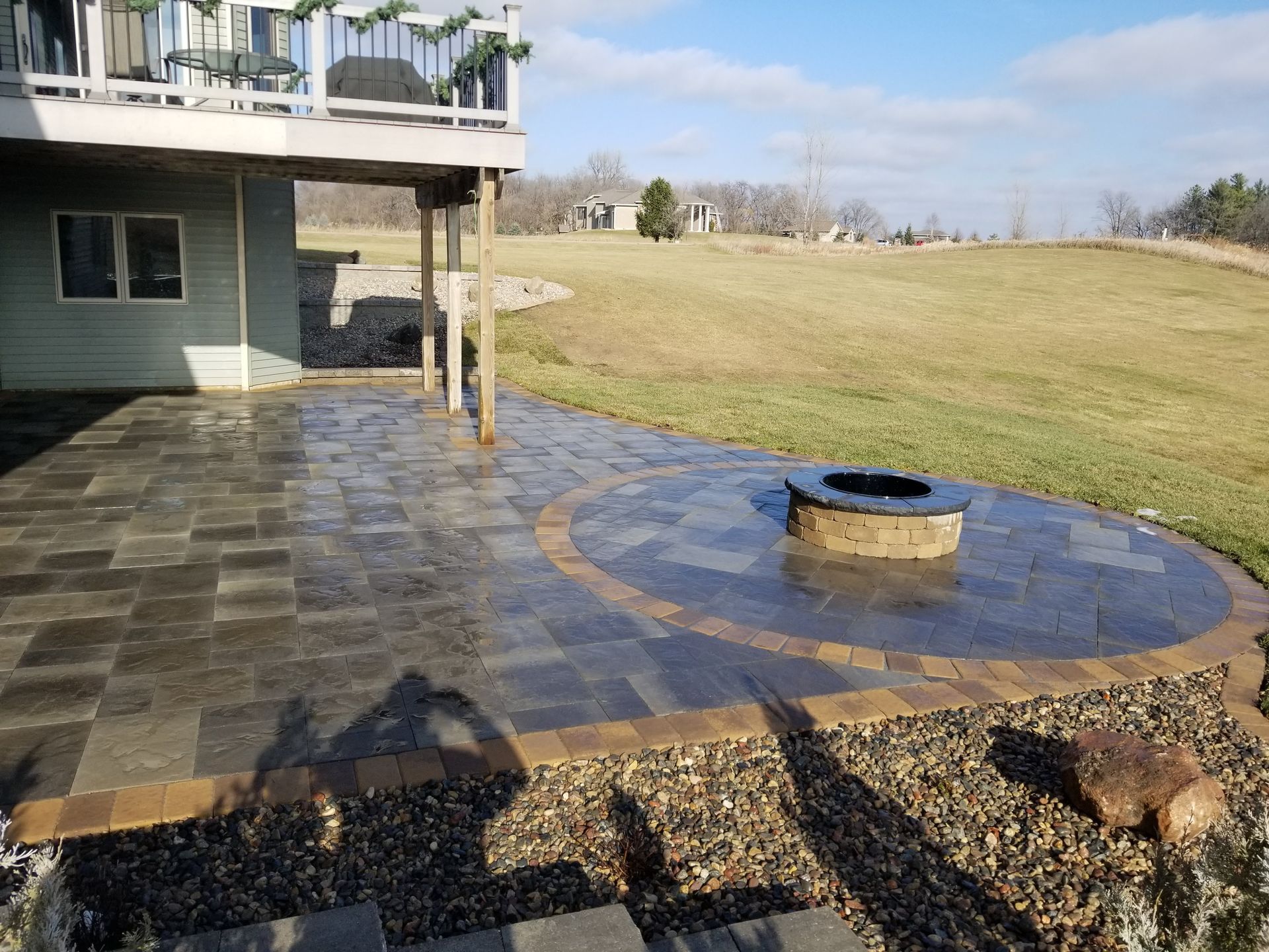 A modern patio with dark stone pavers, a circular fire pit, and a grassy backyard under a clear blue sky.