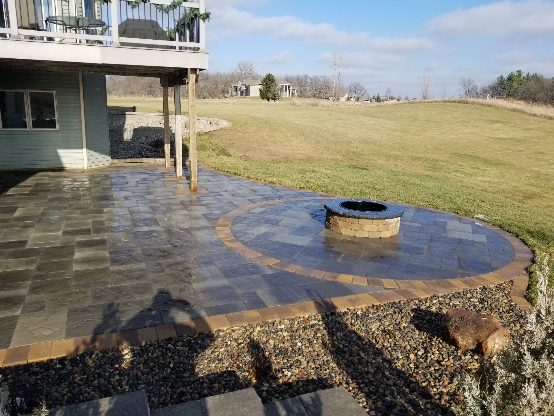 A stone patio with a circular fire pit feature, located under a wooden deck in a grassy backyard setting.