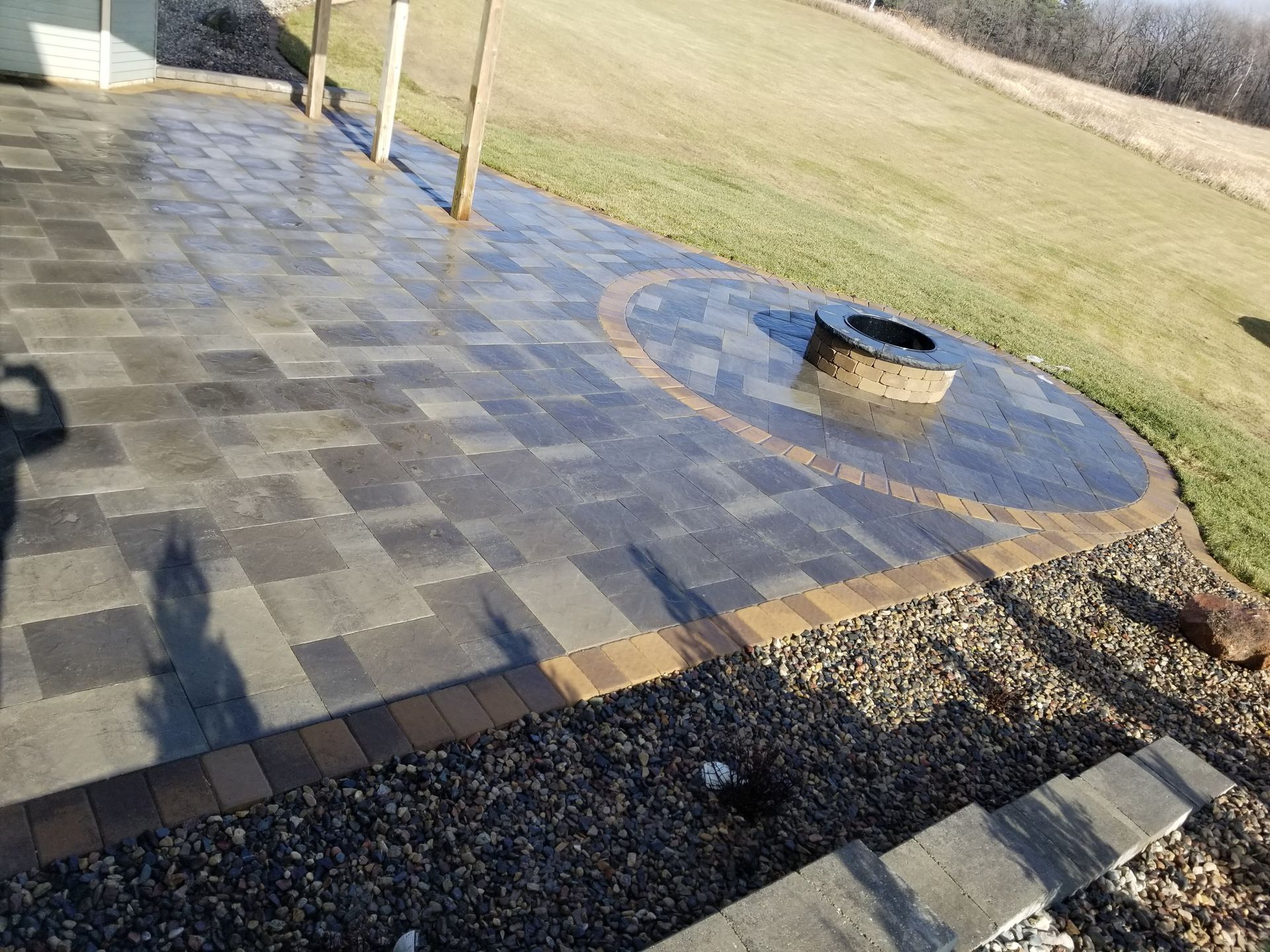 A gray stone patio with a circular fire pit and border trim, viewed from a slightly elevated outdoor setting.