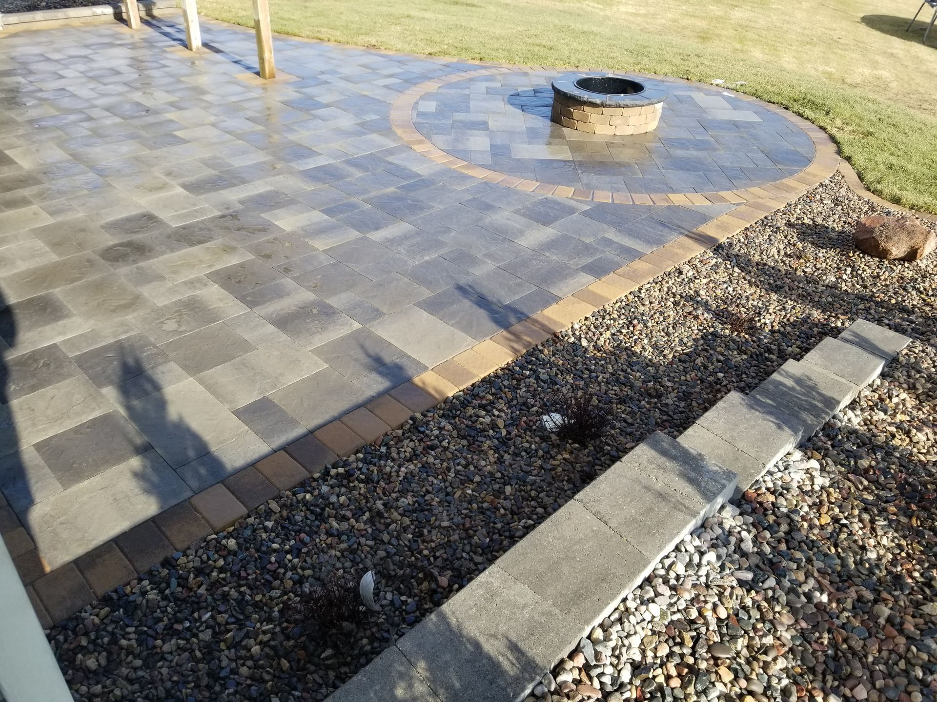 A stone patio with a circular fire pit, bordered by a tan paver edge, adjacent to gravel, a short retaining wall, and lawn.