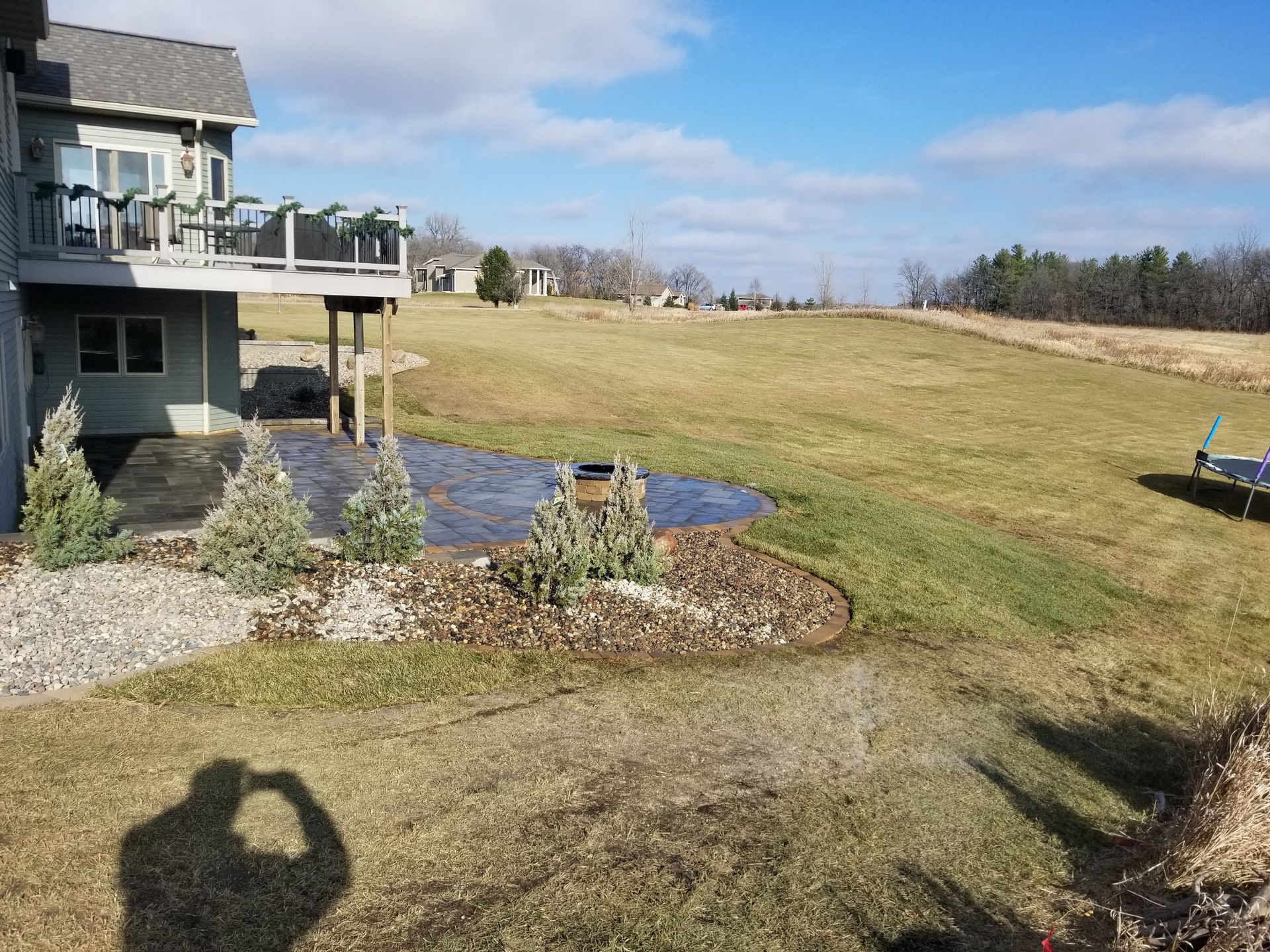 A two-story house with a backyard patio, fire pit, landscaping, and a grassy field under a partly cloudy blue sky.