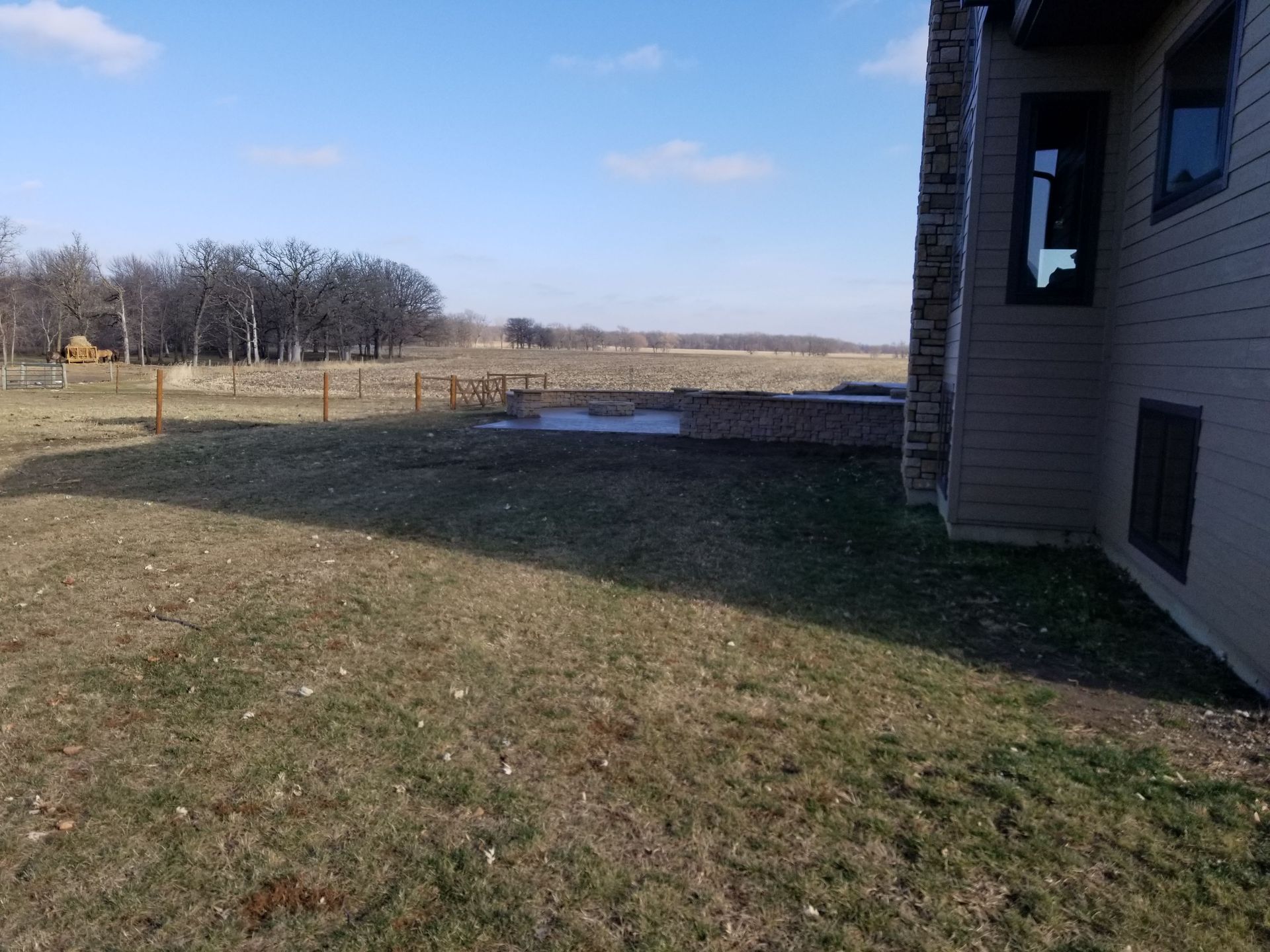 A view of a backyard featuring a grass lawn, a patio, and a stone wall next to the side of a house under a clear sky.