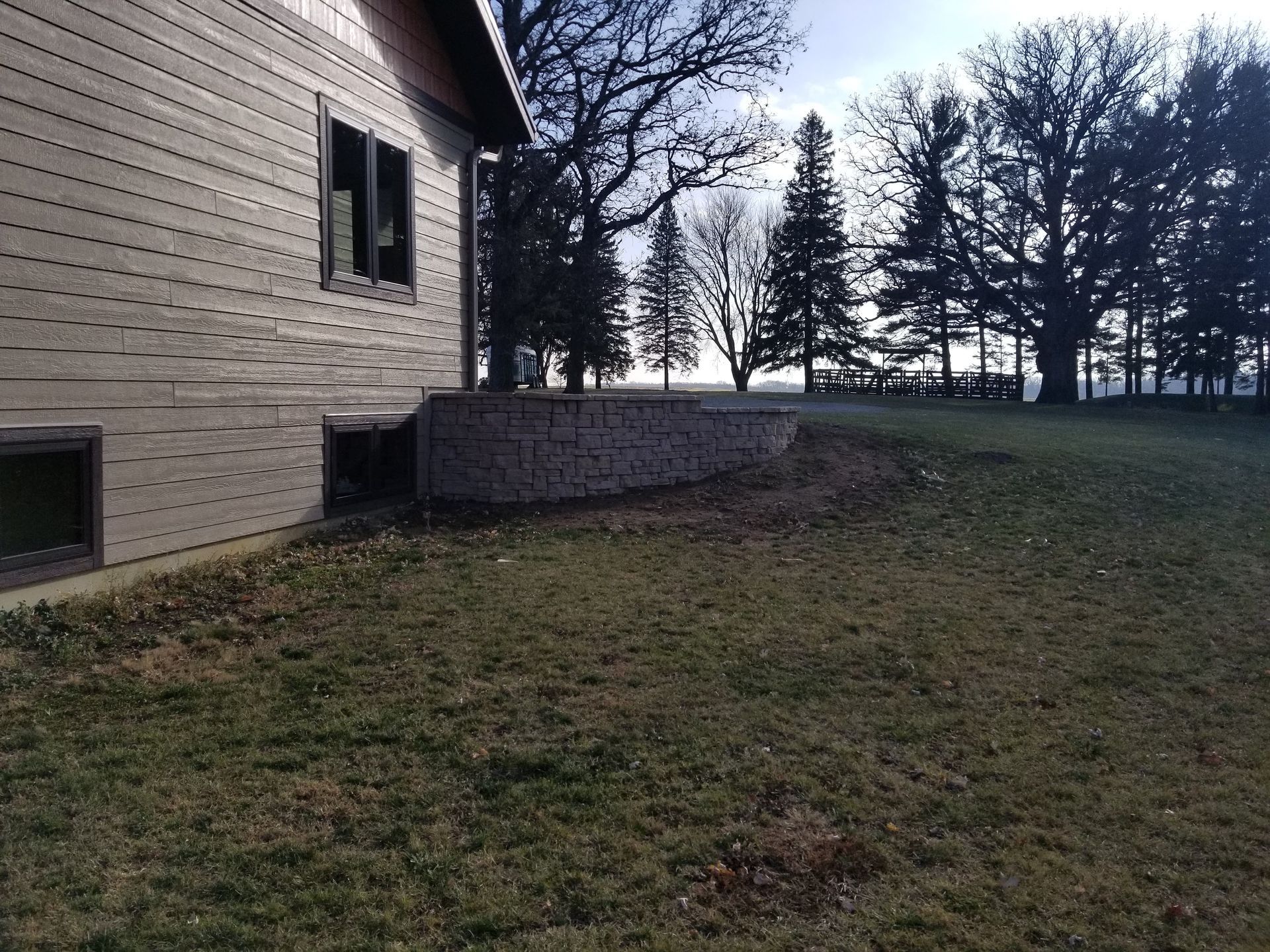 A side view of a tan house with a stone retaining wall in the backyard, surrounded by trees under a sunny sky.