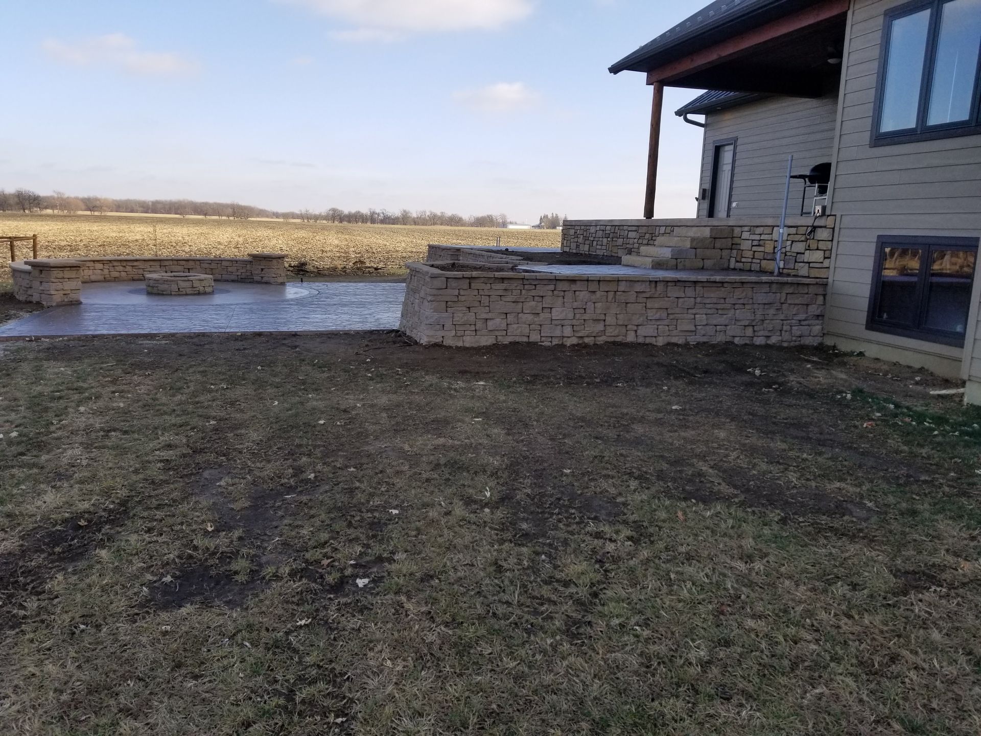 A tan stone patio and retaining wall attached to a light-colored house, overlooking a large field on a sunny day.