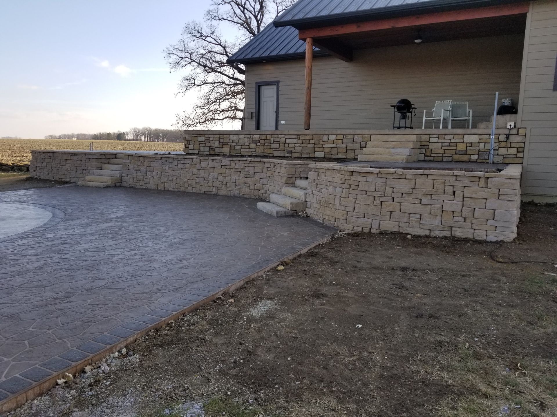 A tan stone retaining wall with integrated steps leading to a house porch and patio in a rural outdoor setting.