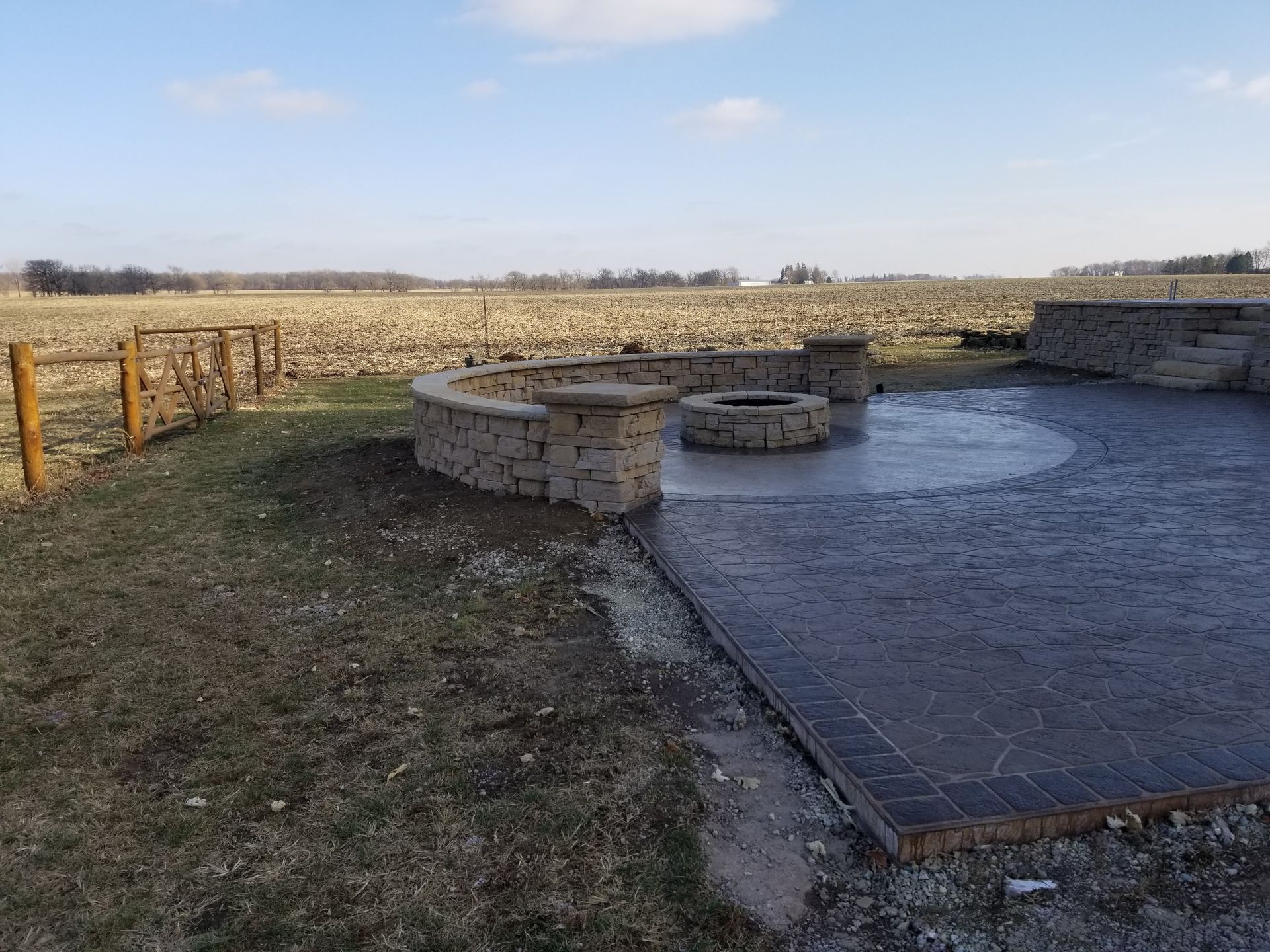 A patio featuring a circular stone fire pit and a curved retaining wall, set against a vast, open field under a blue sky.