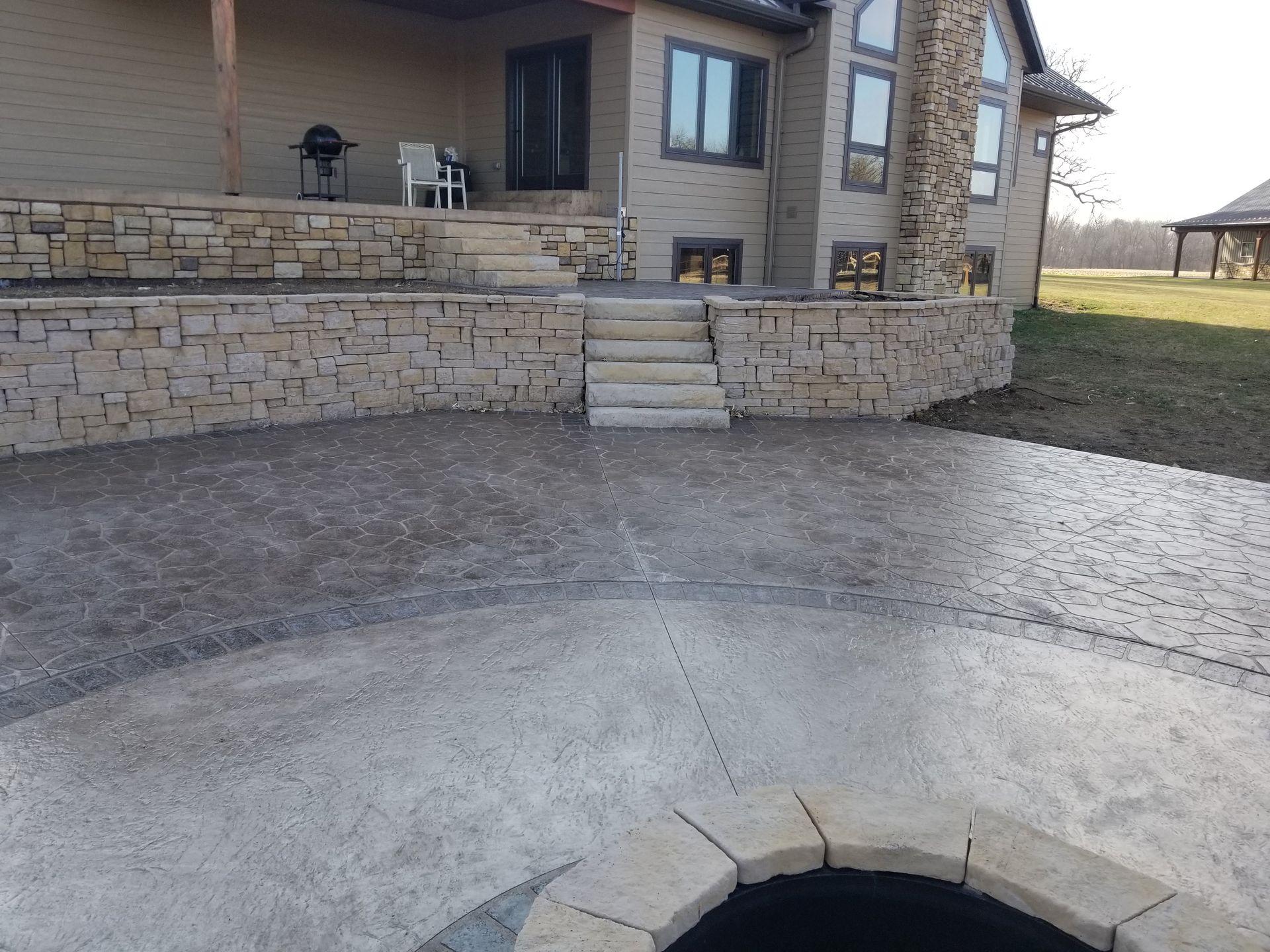 A stone fire pit sits on a stamped concrete patio, featuring tiered stone retaining walls and steps leading to a house.