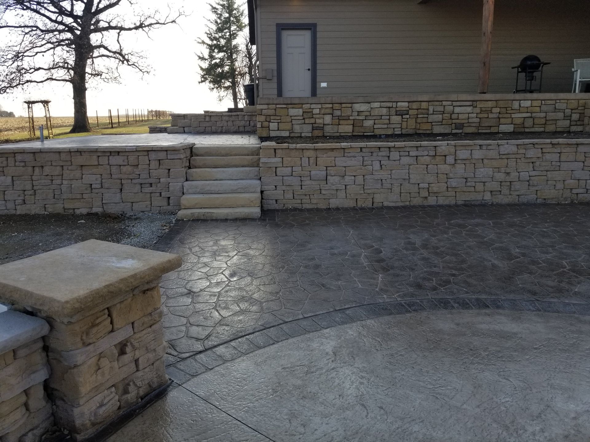 A stone retaining wall with matching stairs leading to a patio area and a house exterior on a sunny day.