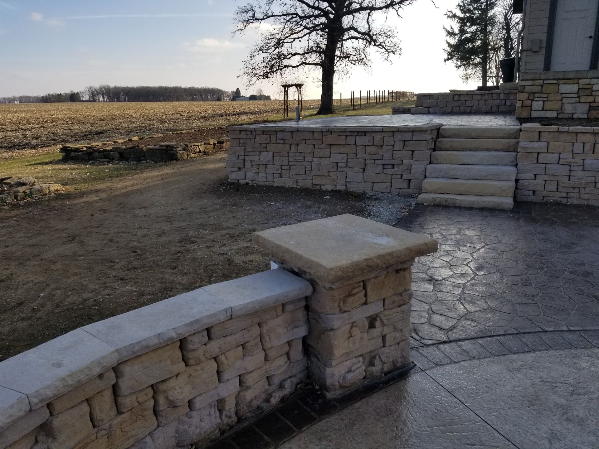 Tan stone wall and matching steps leading to a raised patio with a decorative concrete floor and open field in background.
