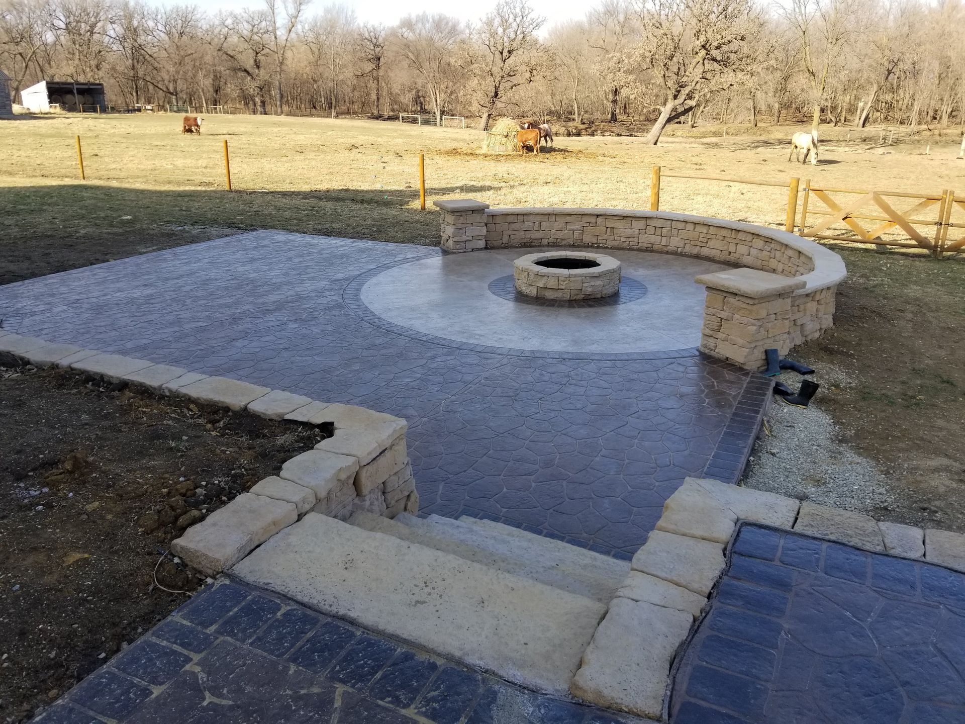 A tiered stamped concrete patio with a circular fire pit and curved stone seating wall, overlooking a rural landscape.