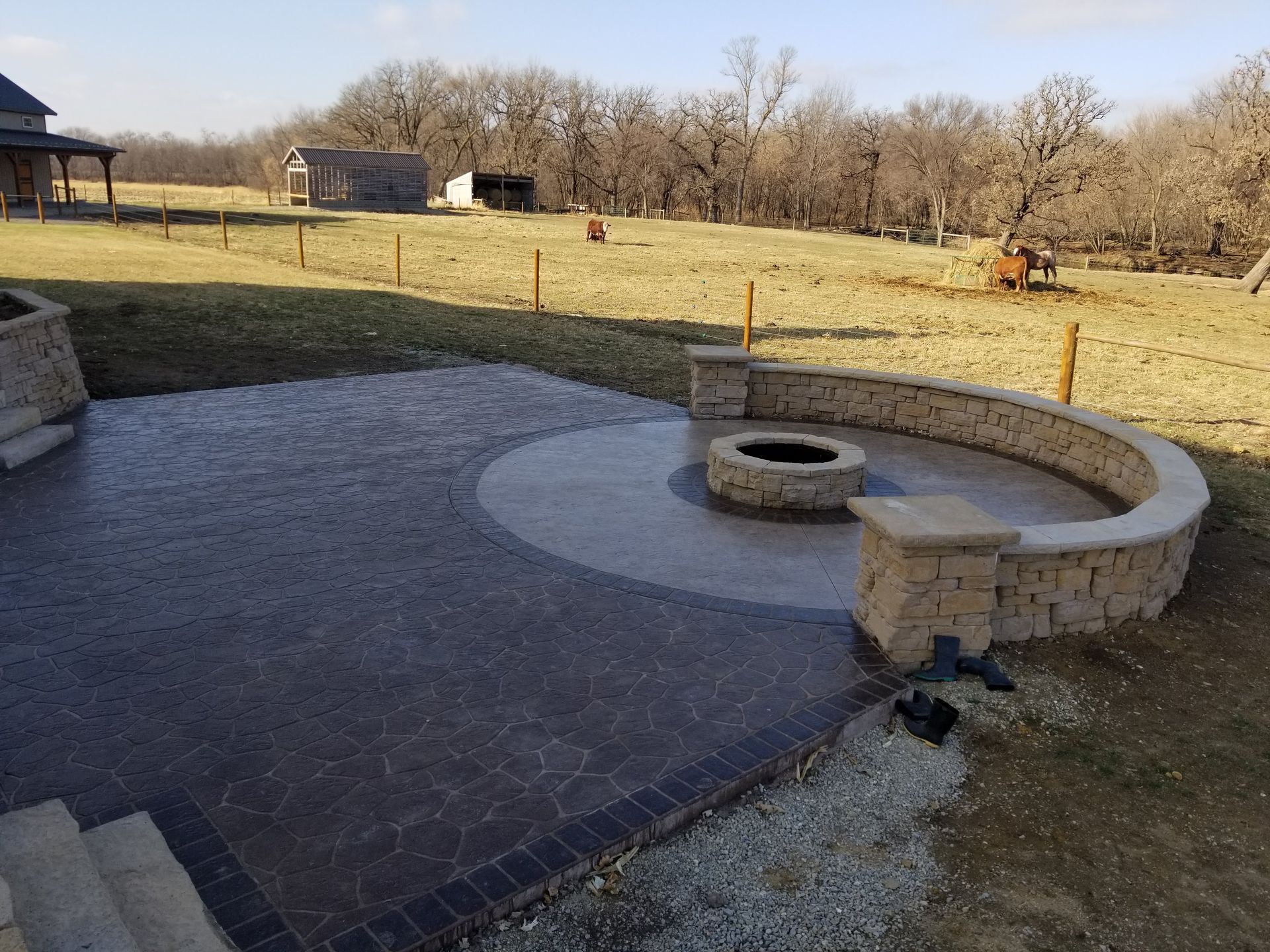 A patterned concrete patio with a circular fire pit and curved stone seating wall in a rural backyard setting.