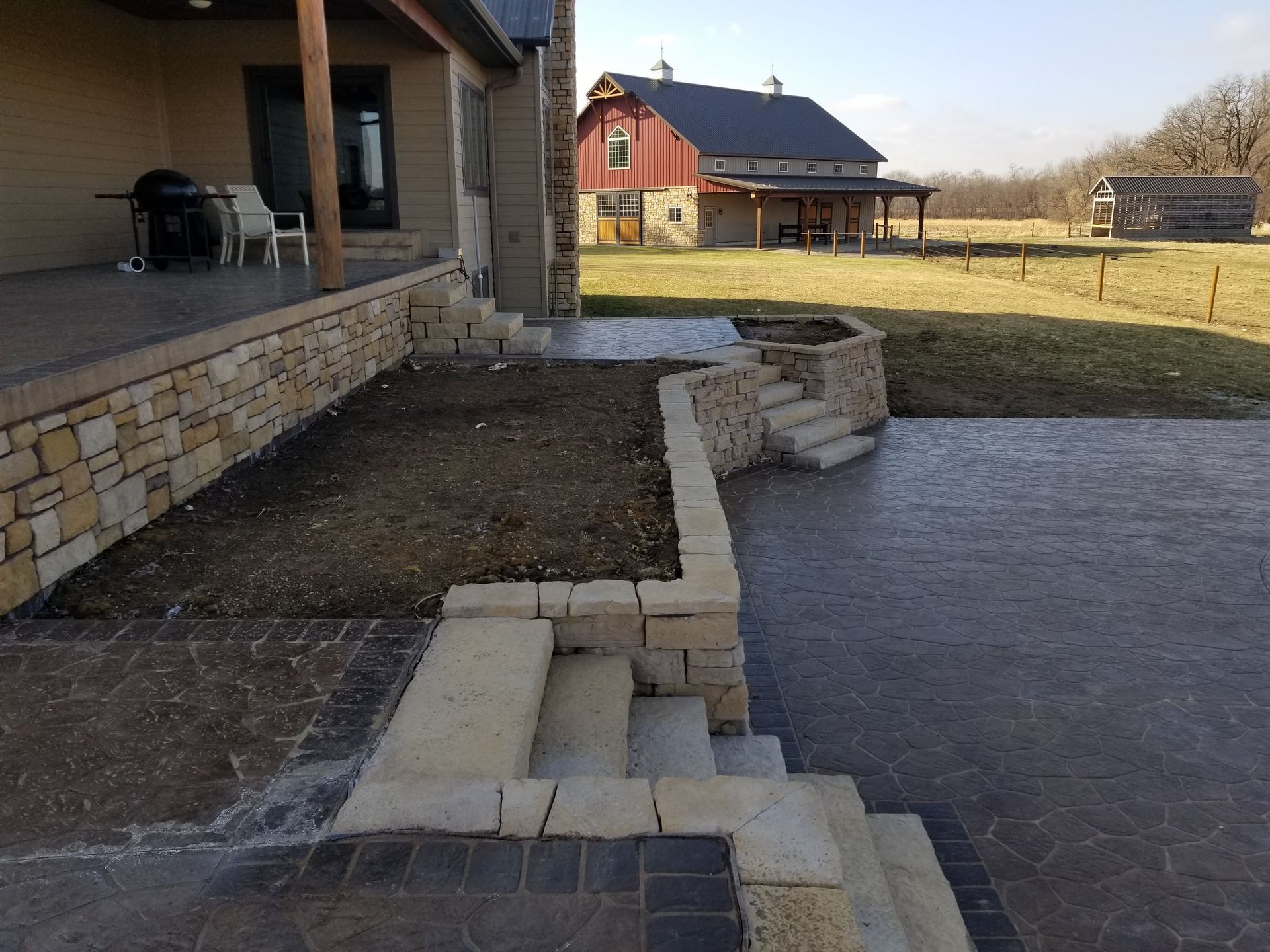 A residential outdoor patio with stone retaining walls and stairs, with a red barn in the distance.