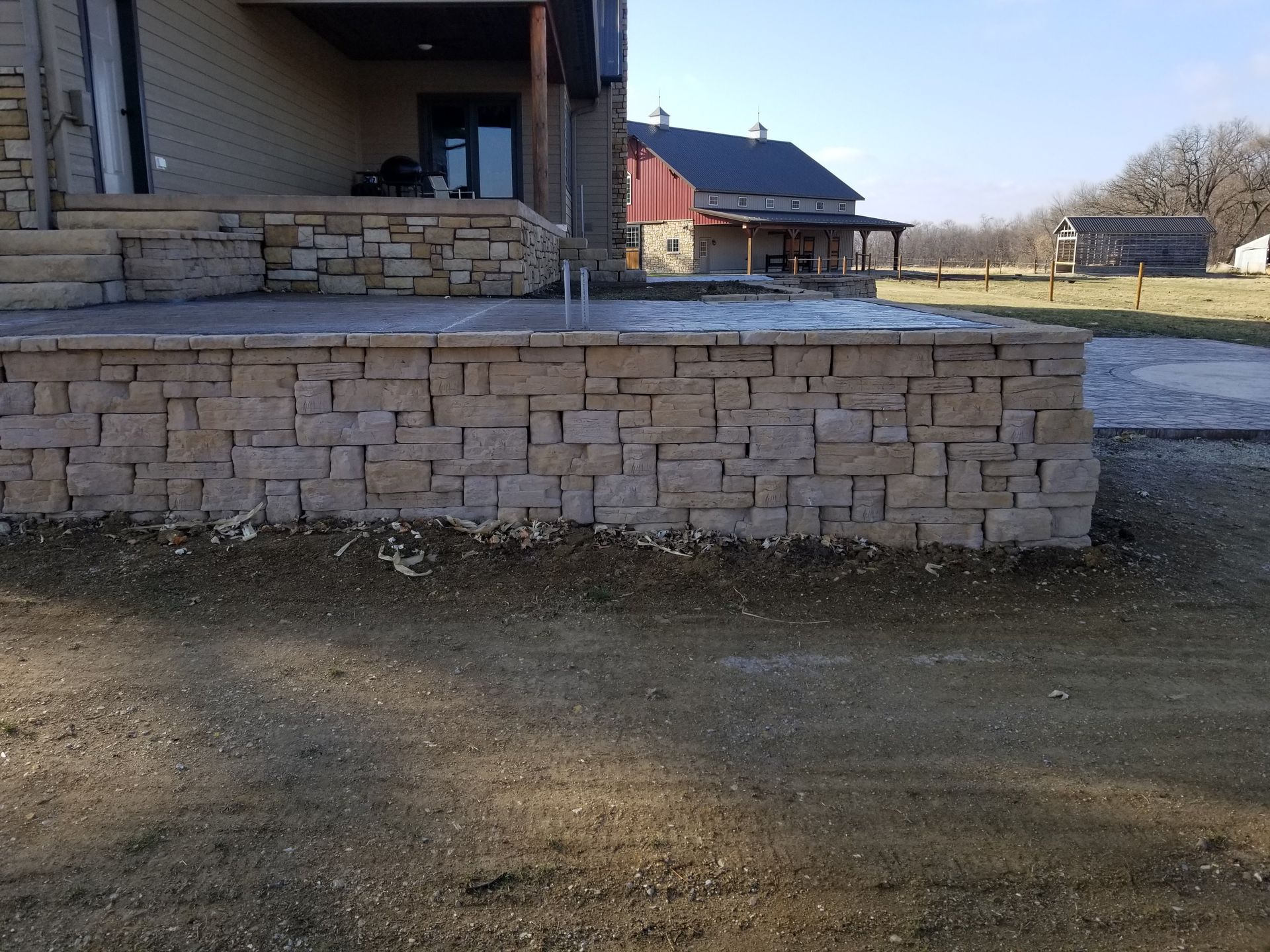A stone retaining wall foundation for a patio next to a house with a farm building in the background.