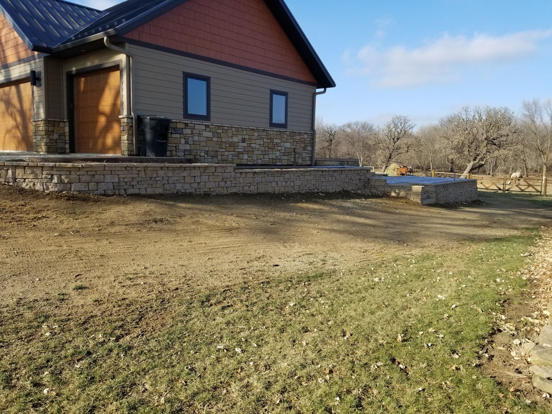A side view of a house with stone siding, a metal roof, and an unfinished landscape featuring a retaining wall.