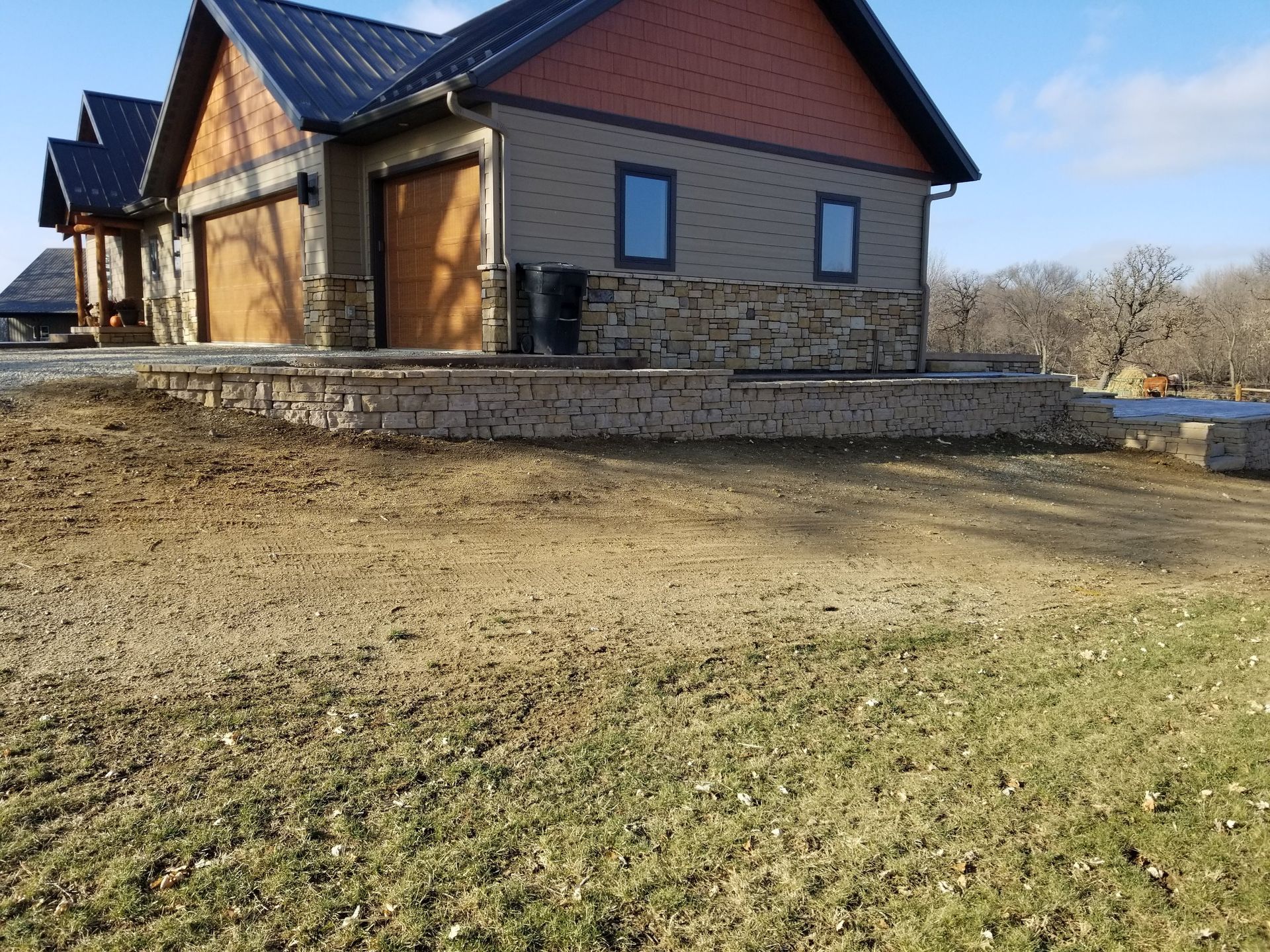 A modern house with a metal roof, tan siding, and stone accents, featuring a stone retaining wall in the front yard.