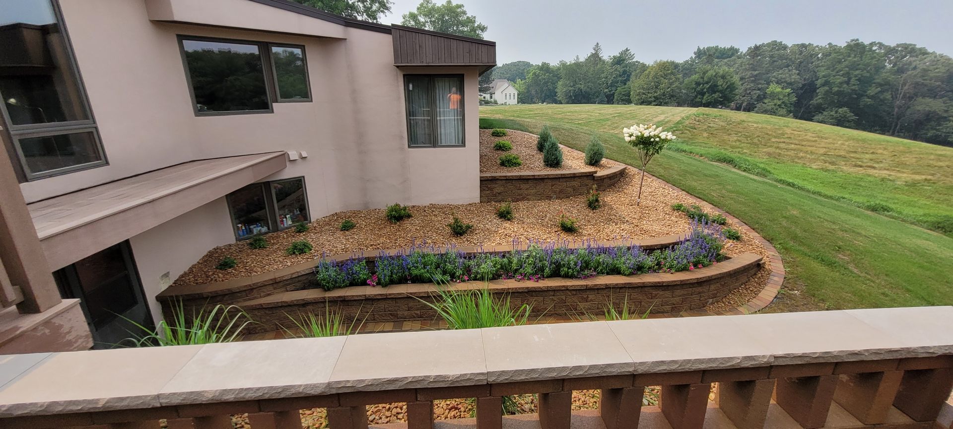 A multi-level tan house with terraced landscaping, stone retaining walls, and purple flowers overlooking a grassy hill.
