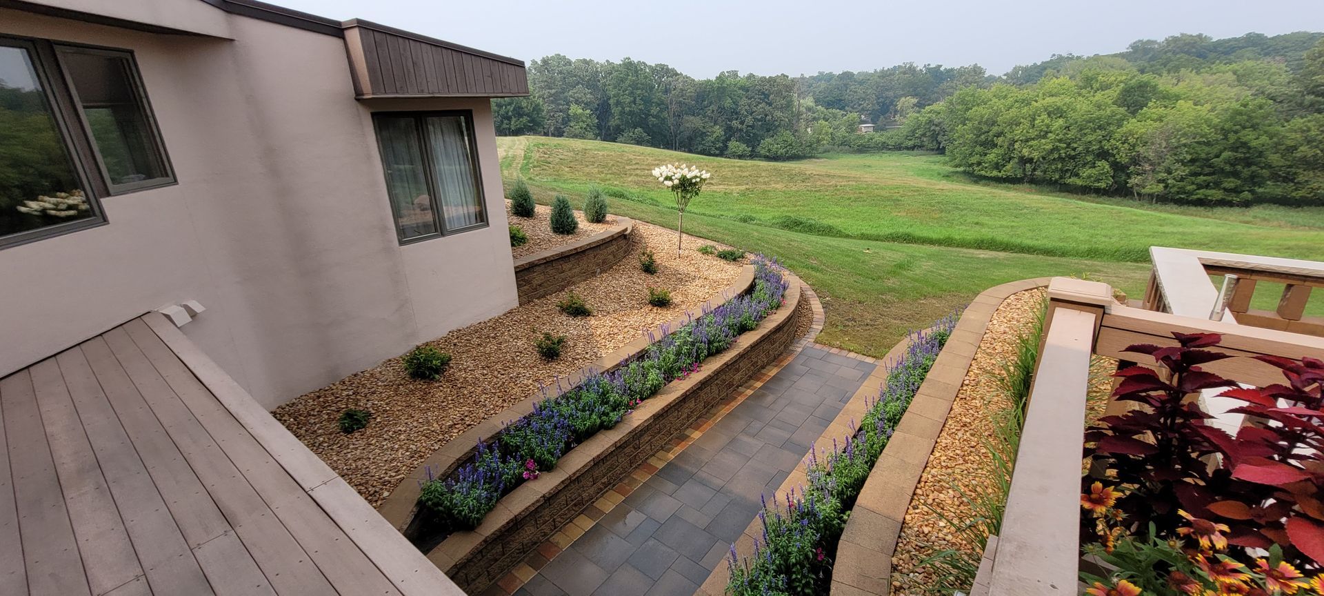 A multi-level terrace with stone walls, gravel, and garden beds leading toward a lush green landscape and open field.