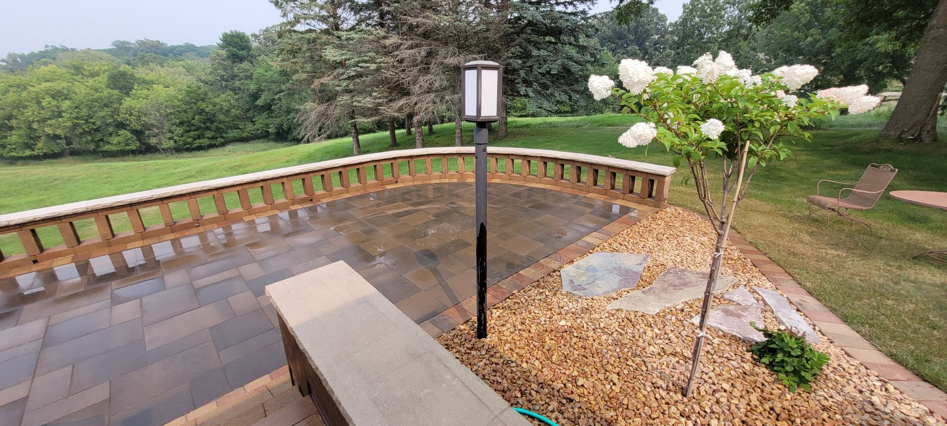 A stone patio with a curved retaining wall, a bench, a light post, and a flowering tree overlooking a grassy landscape.
