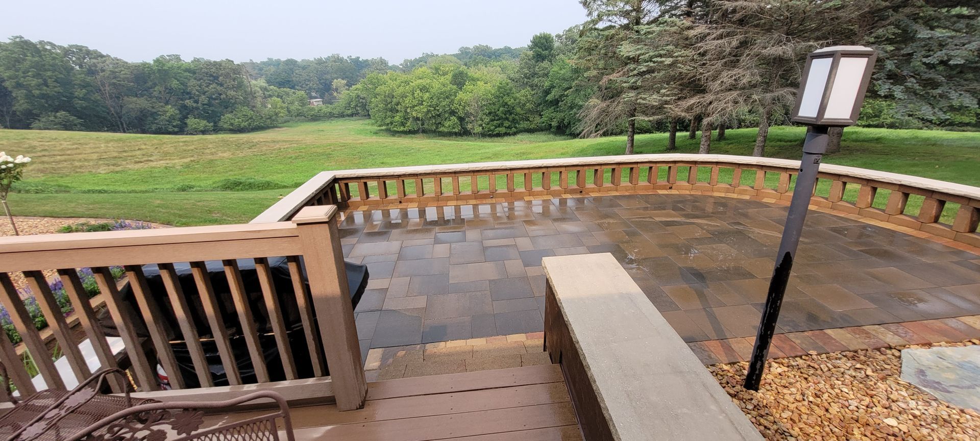 A raised patio with stone pavers, a wooden railing, and a built-in stone bench overlooking a grassy meadow and trees.