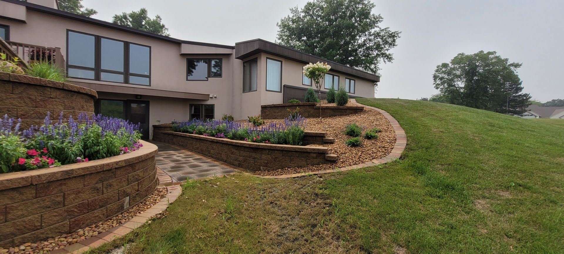 A tan, multi-level house with stone retaining walls, a stone walkway, and landscaped flower beds on a grassy slope.