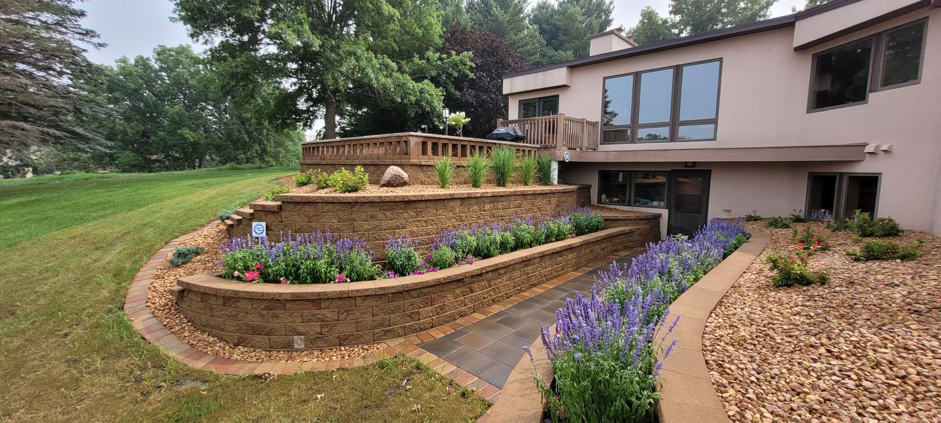 A tiered stone retaining wall landscape with purple flowers and a gravel ground cover beside a multi-level house.