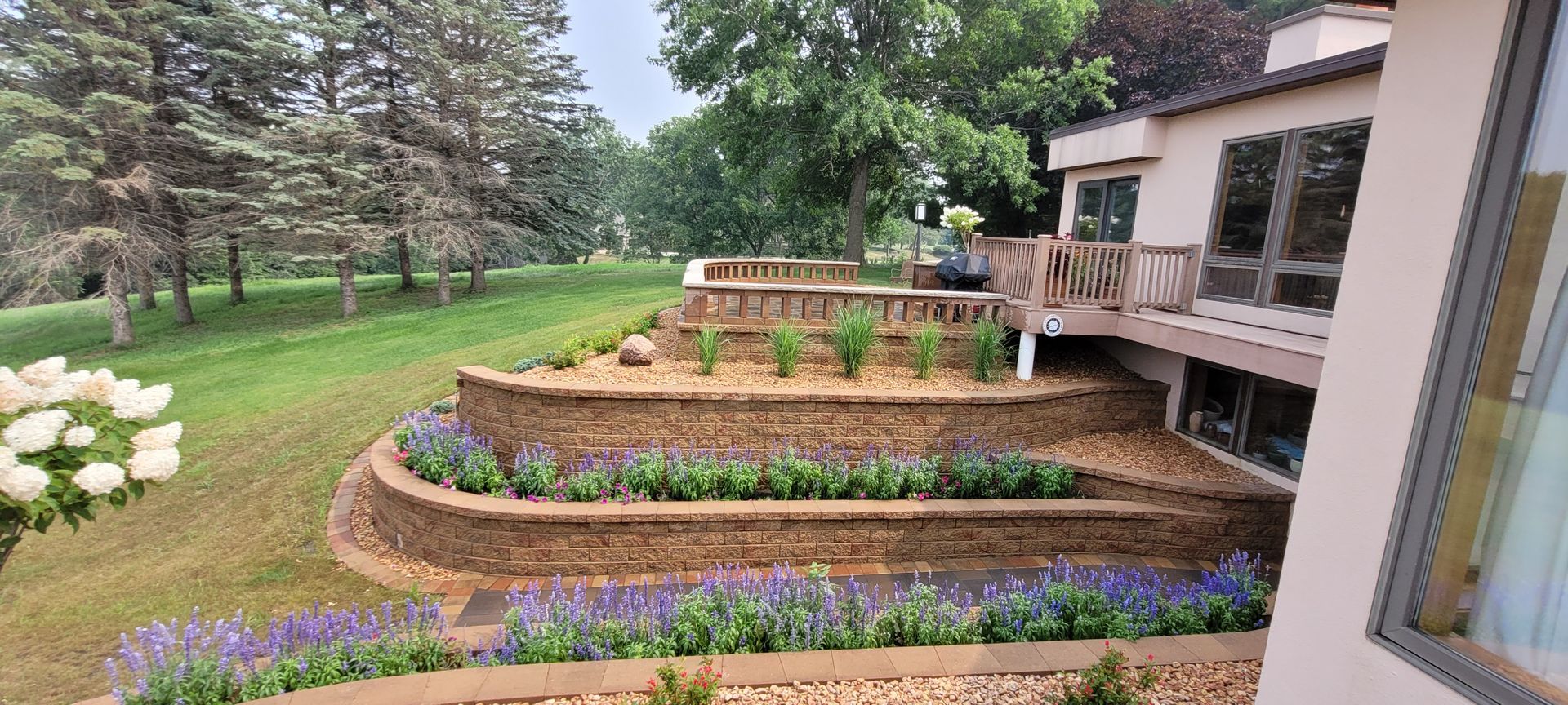 Terraced stone retaining walls landscaped with purple and green plants, alongside a house with a wooden deck.