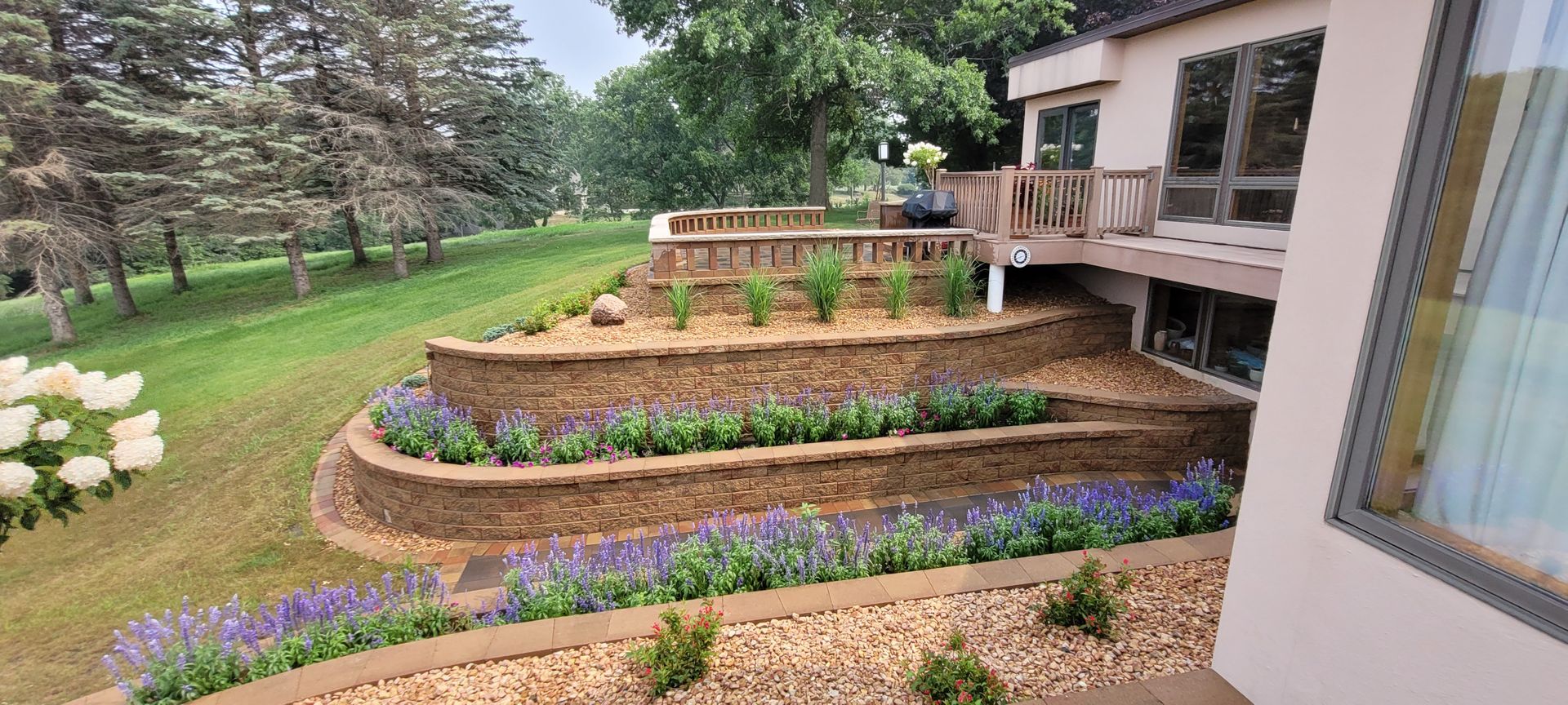 A multi-level backyard landscape with stone retaining walls, garden beds with blooming flowers, and a wooden deck.