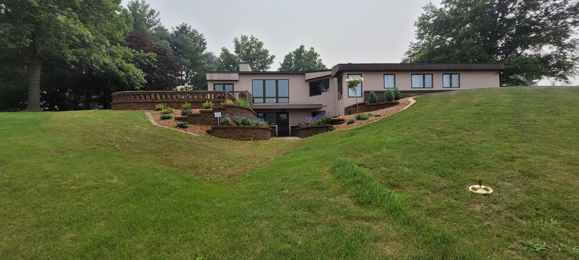 A single-story, light-colored house with a deck and tiered landscaping, set against a backdrop of trees.