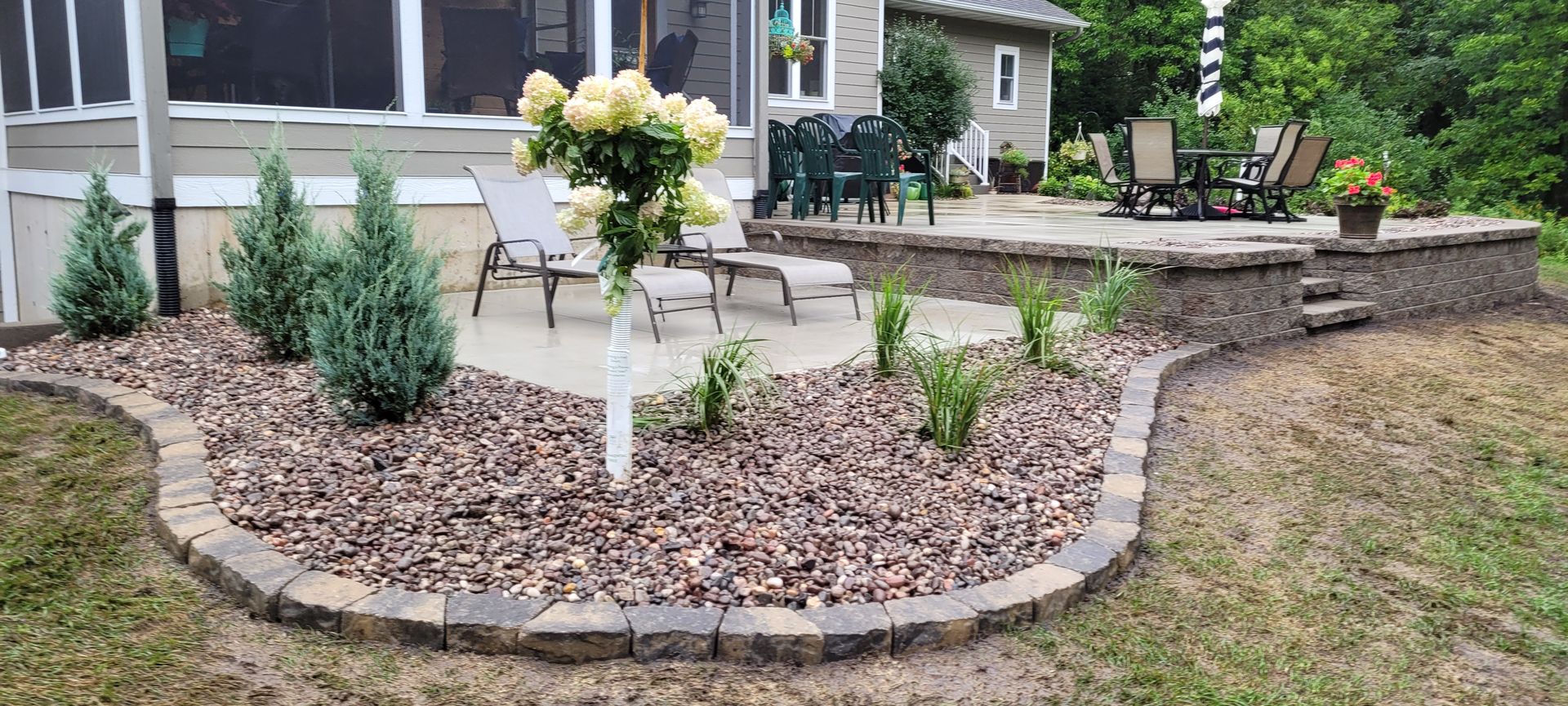 A landscaped patio area featuring a stone border, rock mulch, several small evergreen shrubs, and outdoor furniture.