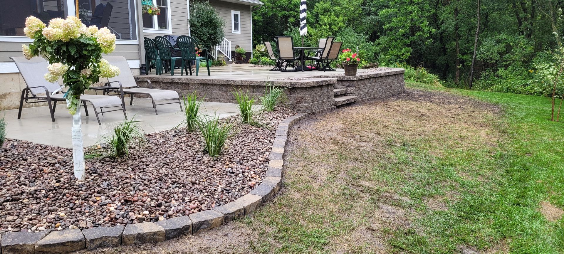 A stone-edged rock garden with flowering shrubs sits beside a paved patio with lounge chairs and a dining set.