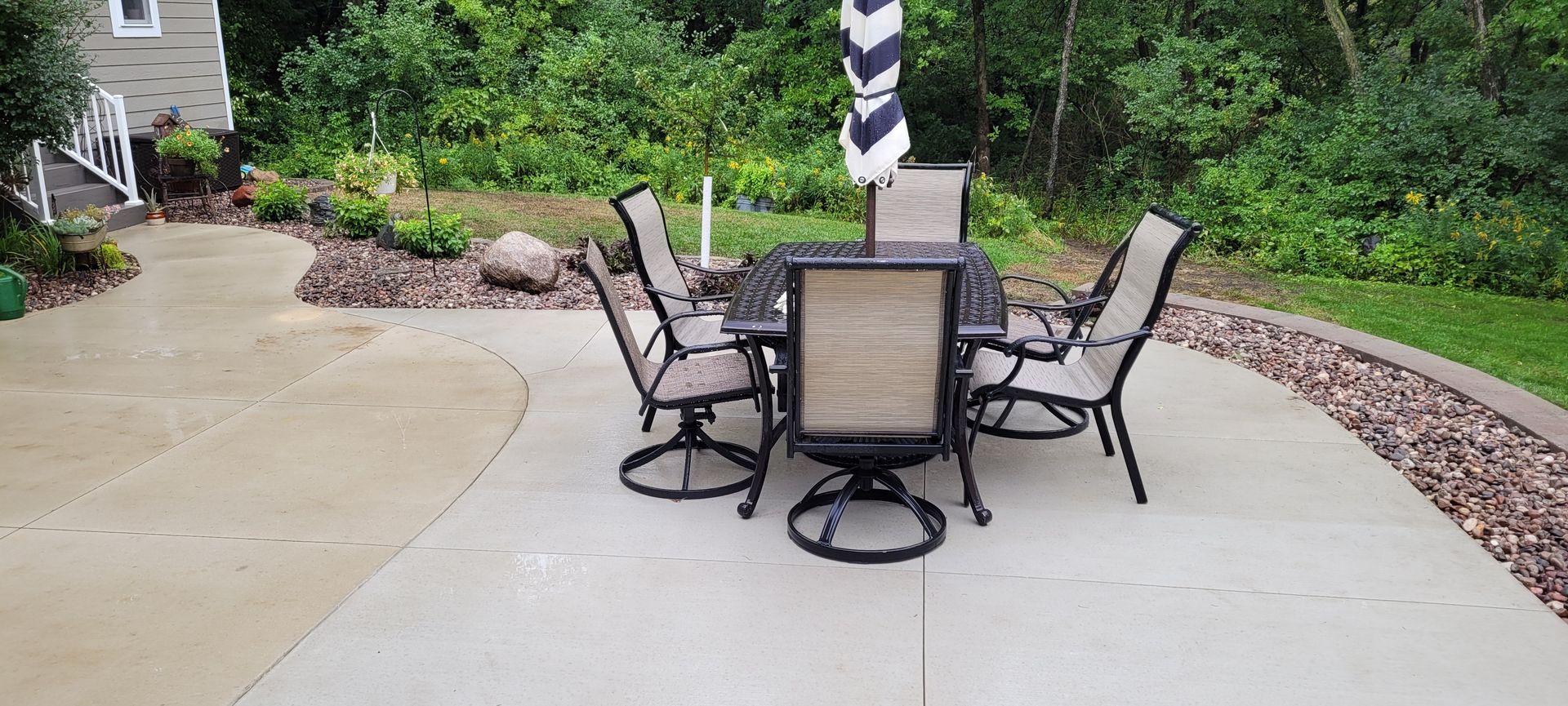 A patio set with four tan chairs and a patterned umbrella on a concrete slab in a grassy backyard.
