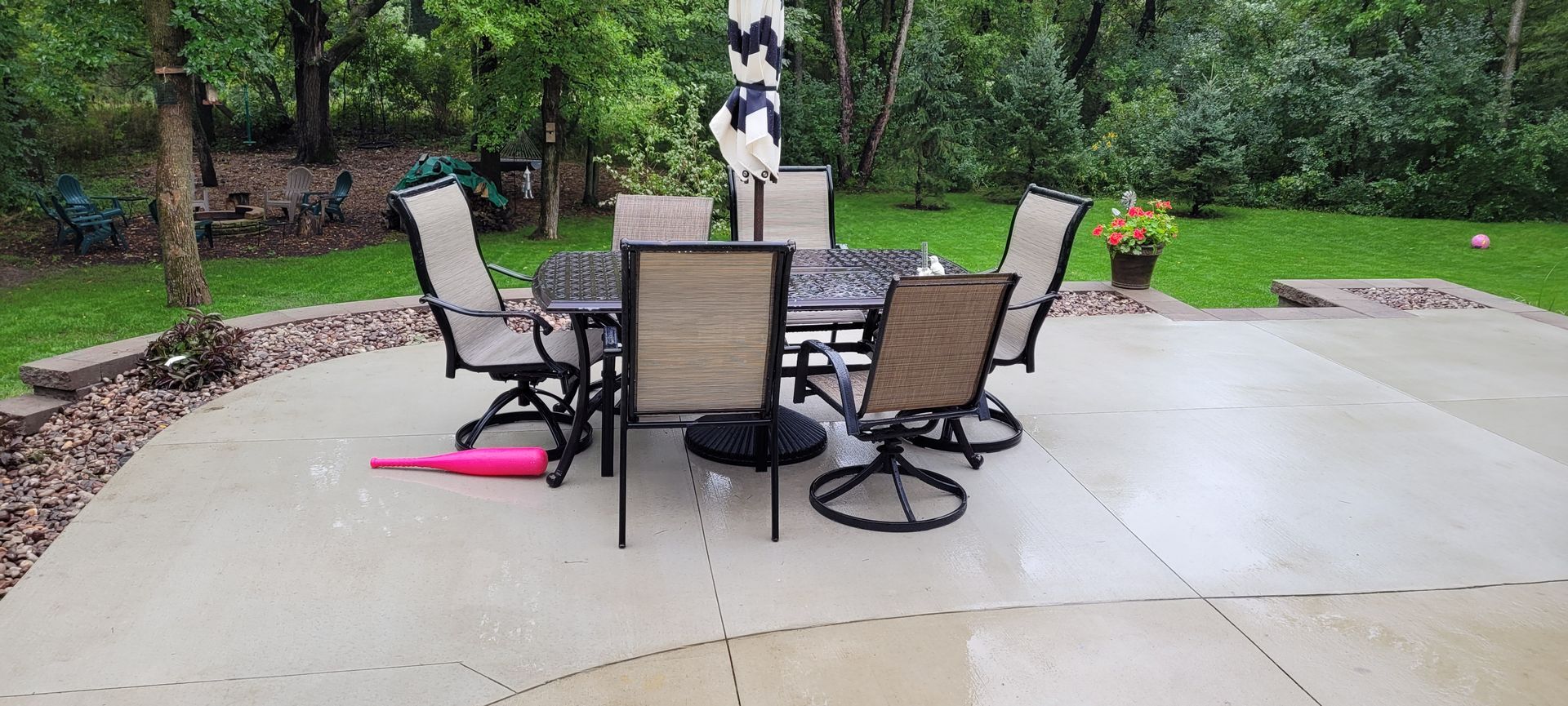 A patio table with four tan chairs and a closed umbrella on a concrete slab in a grassy backyard.