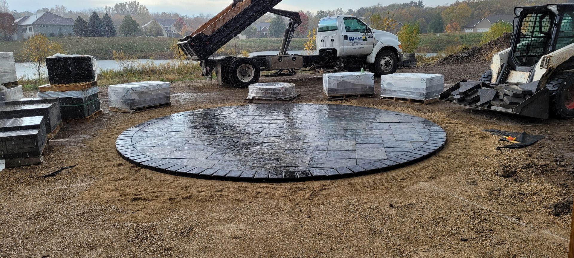 A circular stone patio base is under construction at a job site with a dump truck and skid steer loader nearby.