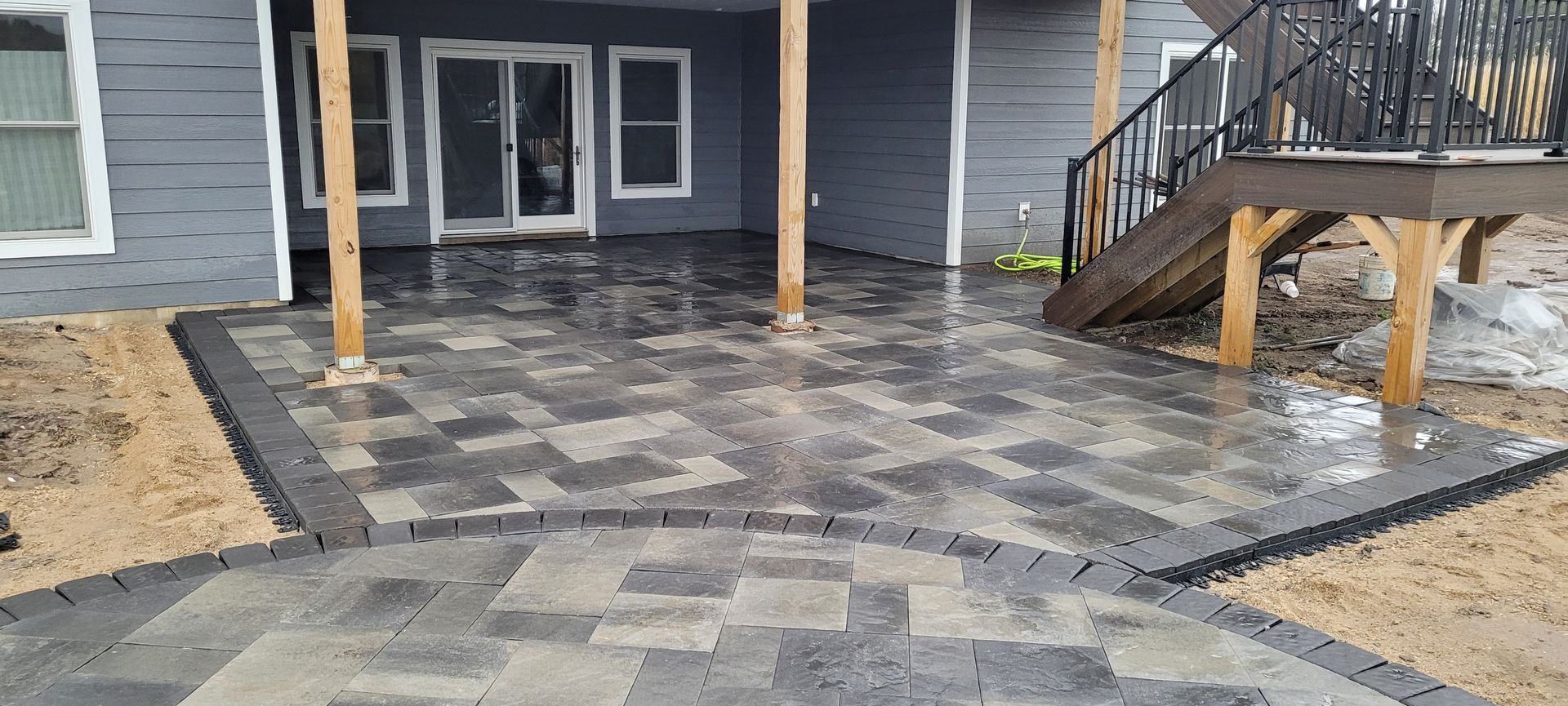 A grey paver patio with a curved border design sits behind a house, with wooden support posts and a deck staircase.