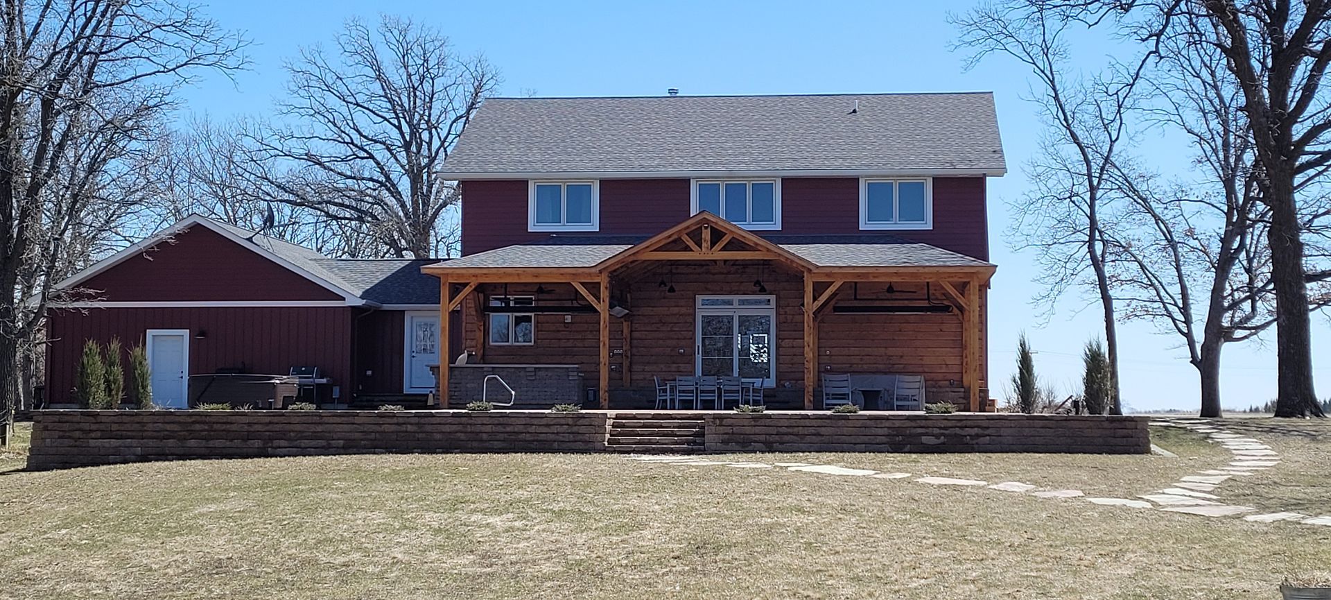 A two-story red farmhouse with a wooden patio cover and stone retaining wall sits on a grassy field under a blue sky.