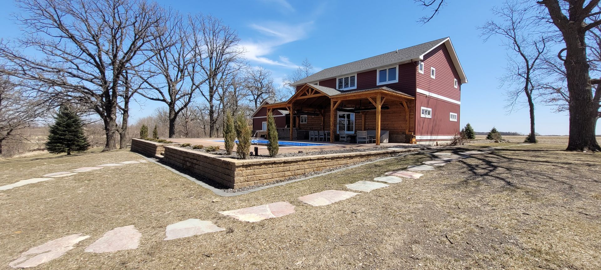 A two-story red farmhouse with a stone patio, wooden pergola, and pool, surrounded by bare trees under a clear blue sky.
