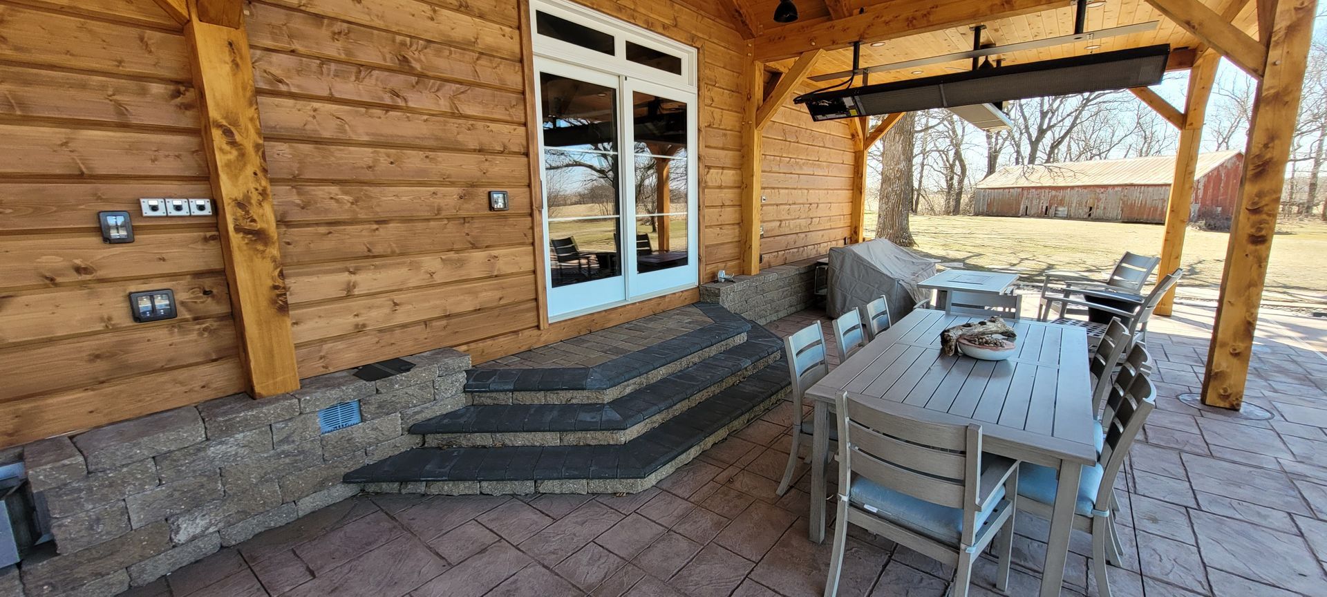 A wooden log cabin patio features a gray stone staircase leading to glass doors, a dining set, and an outdoor heater.