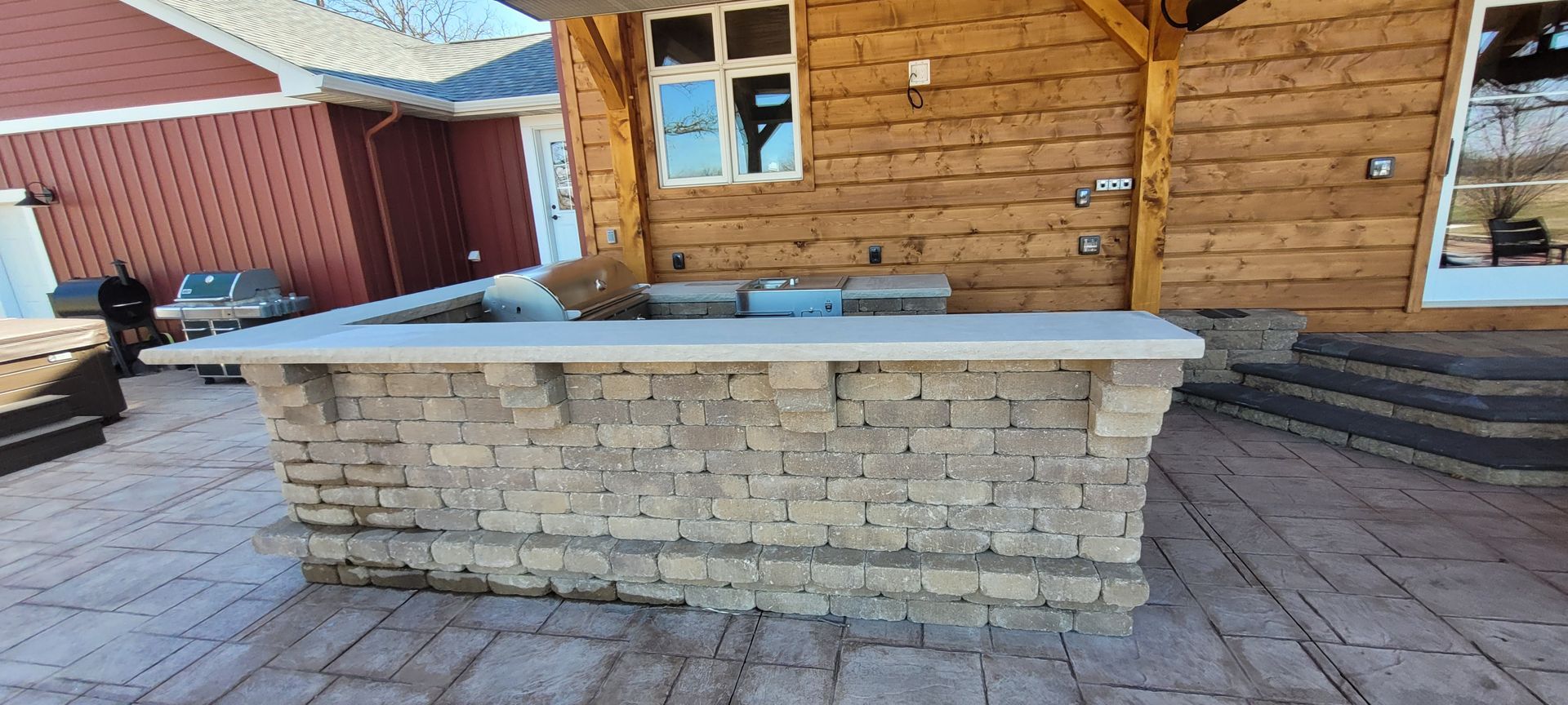 Outdoor kitchen bar with light stone base and a smooth concrete countertop, set on a patio near a log cabin wall.