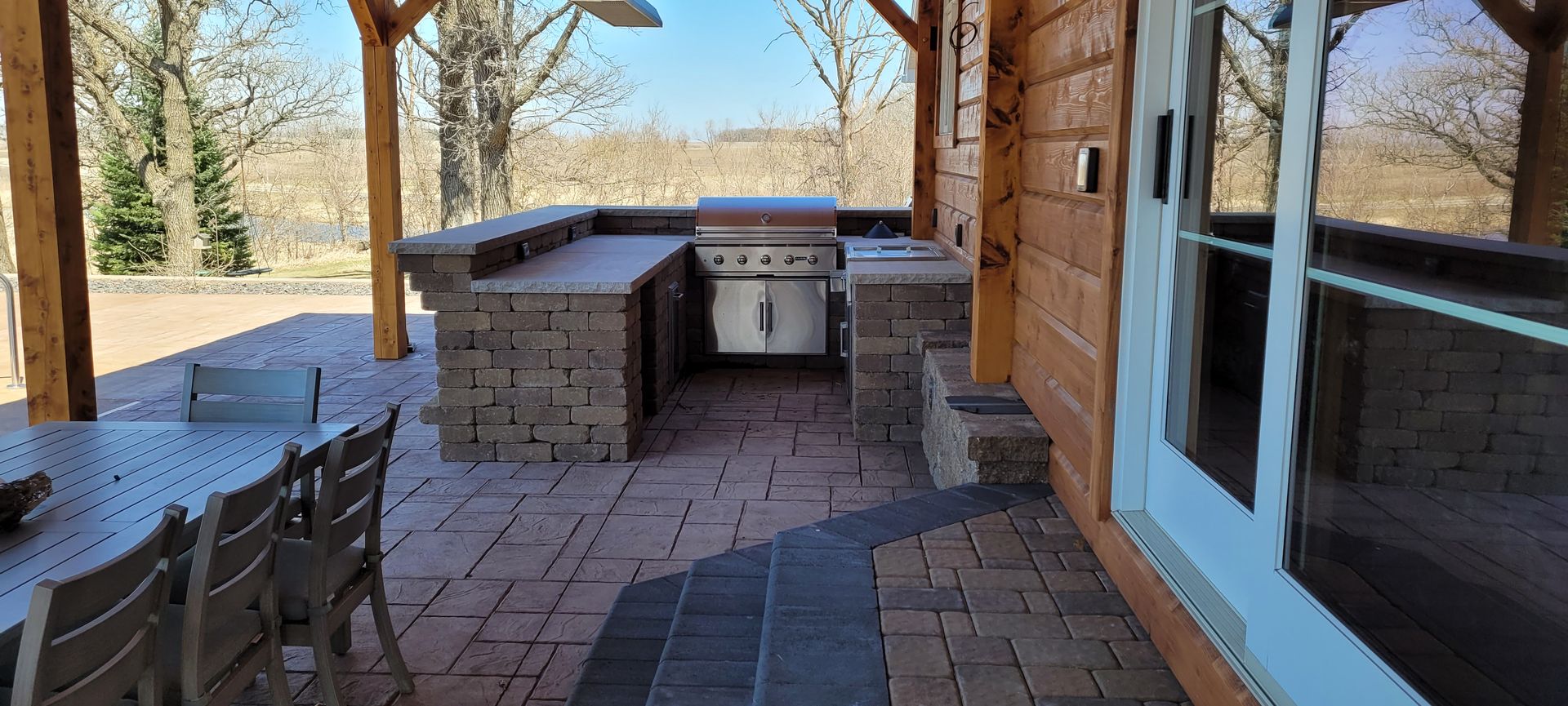 A stone outdoor kitchen with a built-in grill and counter on a brick patio under a covered wooden patio structure.