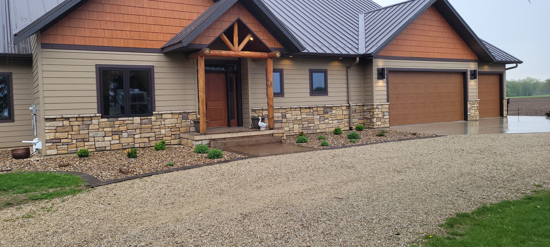 A single-story suburban house with stone accents, horizontal tan siding, brown wooden garage doors, and a gravel driveway.