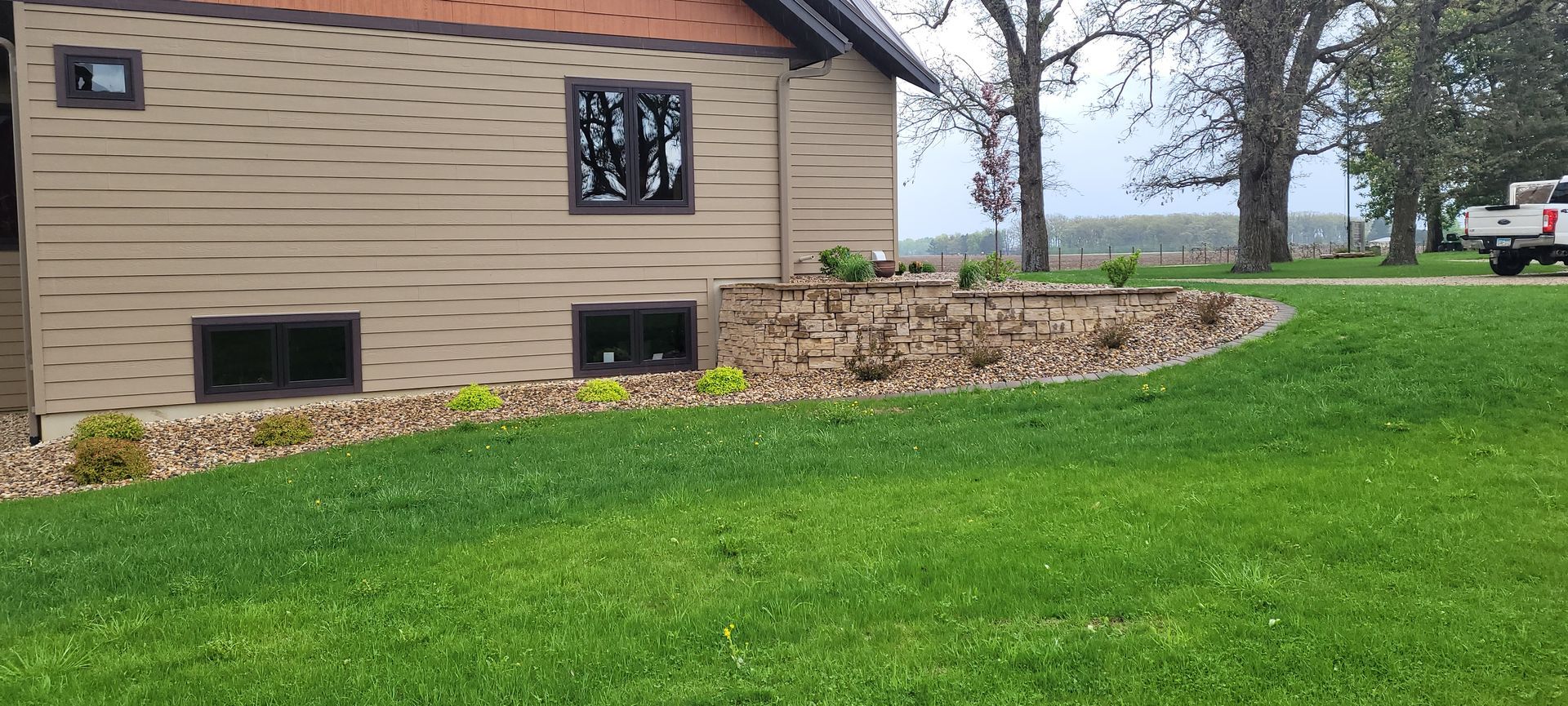 A beige house with dark trim and windows sits beside a landscaped stone garden bed against a green lawn.