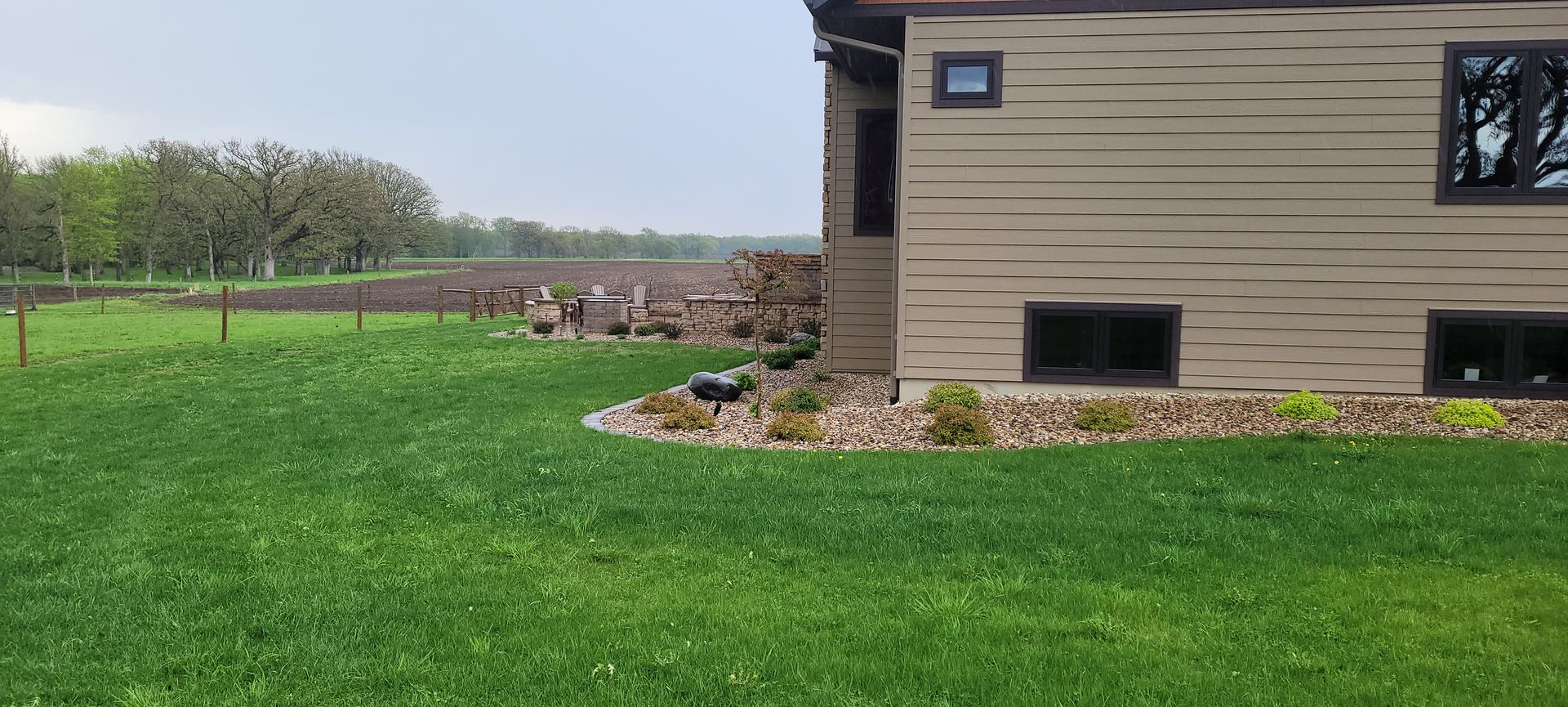 A side view of a tan house with a stone garden bed, adjacent to a green lawn, a fenced field, and a tree line.
