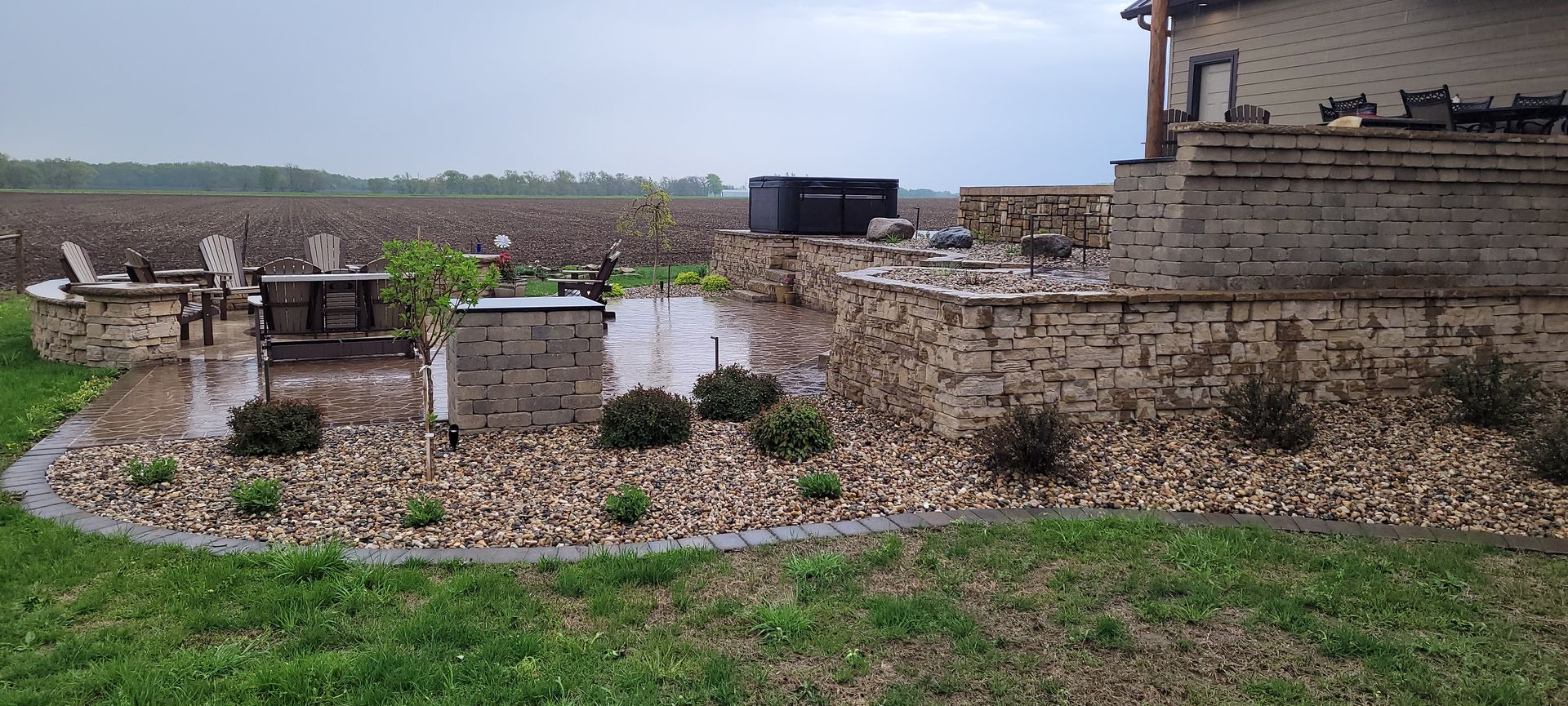 An outdoor patio with stone walls, furniture, and landscaping, overlooking a large field under an overcast sky.