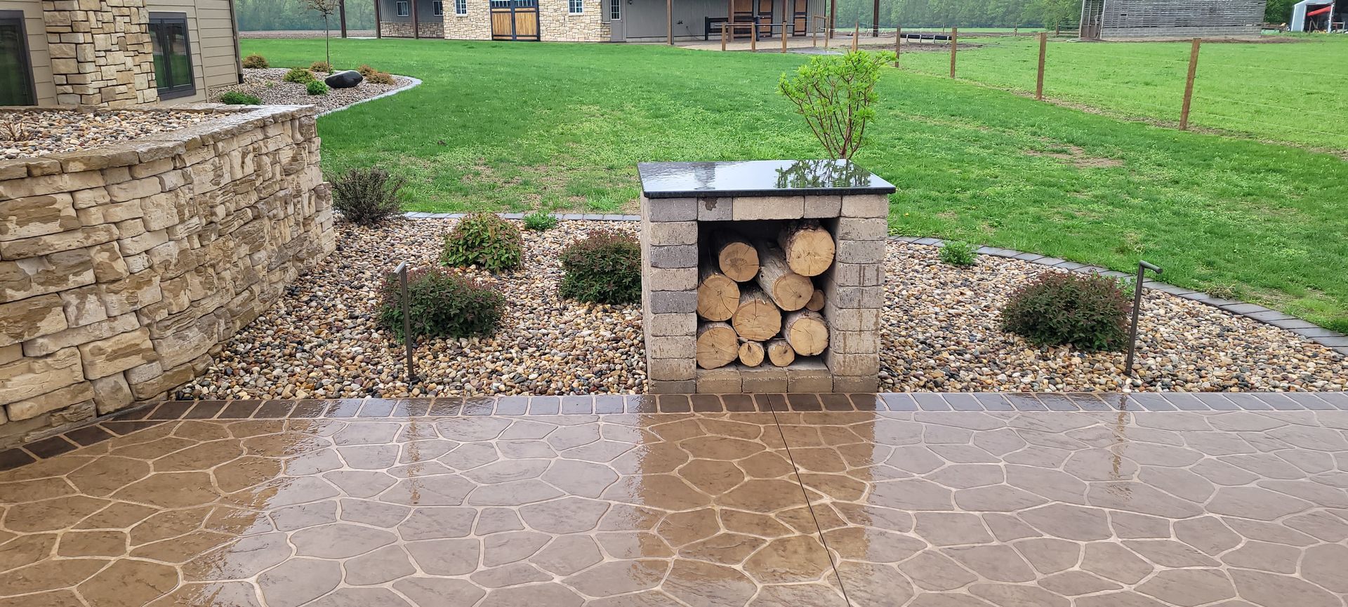 A stone patio with a built-in stone wood-storage unit holding logs, set in a yard with gravel and small bushes during rain.