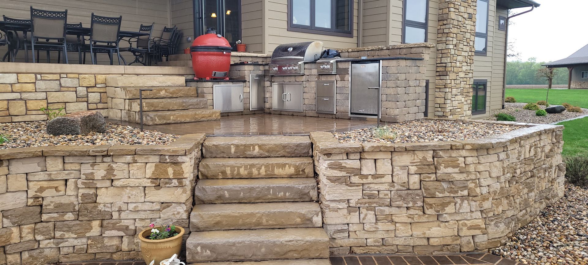 A multi-level stone patio with a built-in outdoor kitchen, stairs, and a red kamado-style grill.