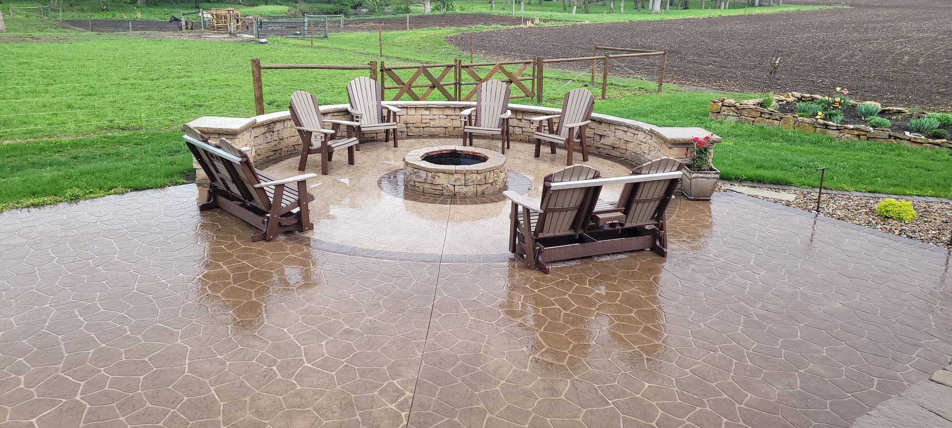 Outdoor patio with stone fire pit and chairs covered in hail during a rainstorm, set against a grassy field and farmland.