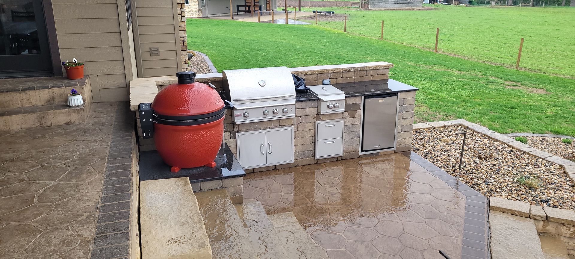 An outdoor kitchen on a patio, featuring a bright red kamado grill, a stainless steel gas grill, and a refrigerator.