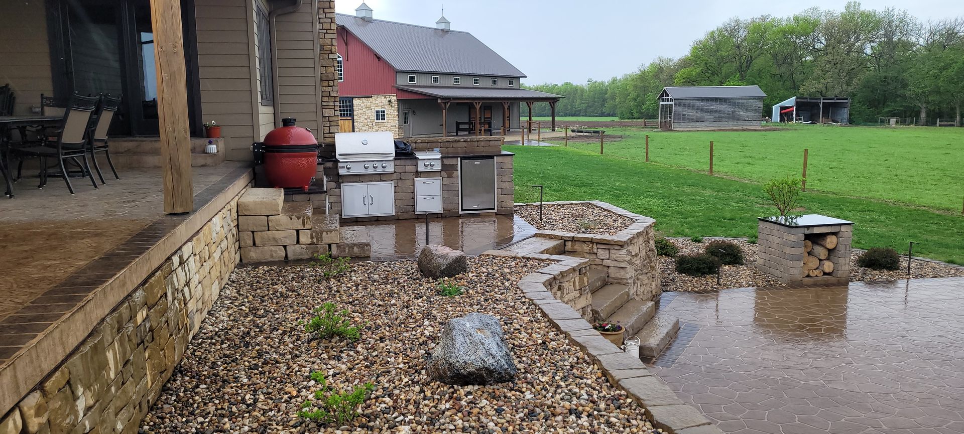 An outdoor patio area with a grill, a red smoker, and stone walls during a rainstorm, overlooking a rural landscape.