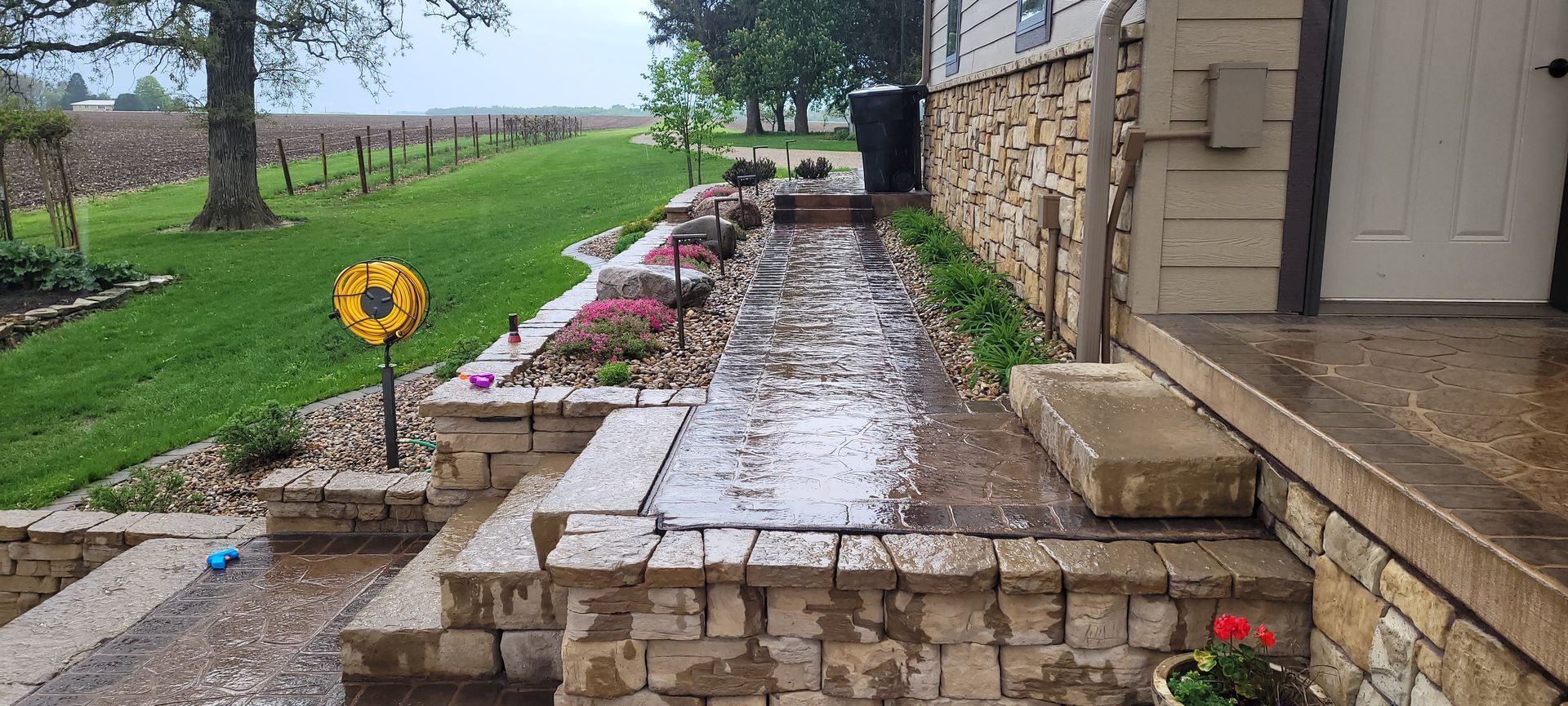 A wet stone walkway and tiered retaining wall border a house exterior with a grassy yard and distant field in the rain.